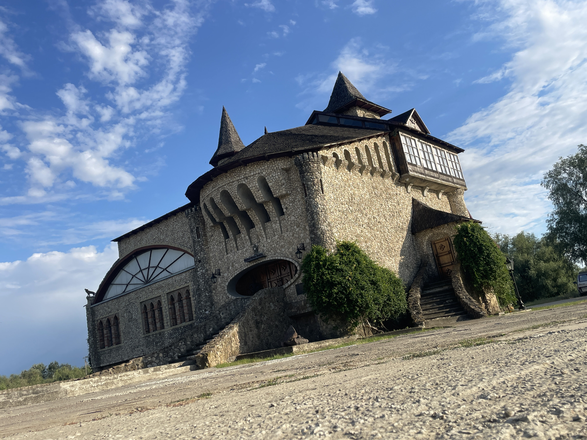 The image shows a distinctive stone building with a medieval, castle-like appearance, located in Colibași, Moldova. The building has multiple pointed roofs covered in dark shingles, reminiscent of turrets. The walls are constructed from irregularly shaped light-colored stones. Arched windows and doorways are visible, some with decorative stonework. A unique feature is a large, arched window with a geometric grid pattern on the left side of the building, and a projecting windowed structure with many panes of glass on the upper right. Several staircases lead up to different entrances, flanked by stone walls and dense green bushes. The ground in front of the building is a rough, sandy or gravelly surface, with some vegetation growing along the edges. The sky is blue with scattered white clouds. Trees are visible in the background, and a portion of a vehicle can be seen on the right side of the image.