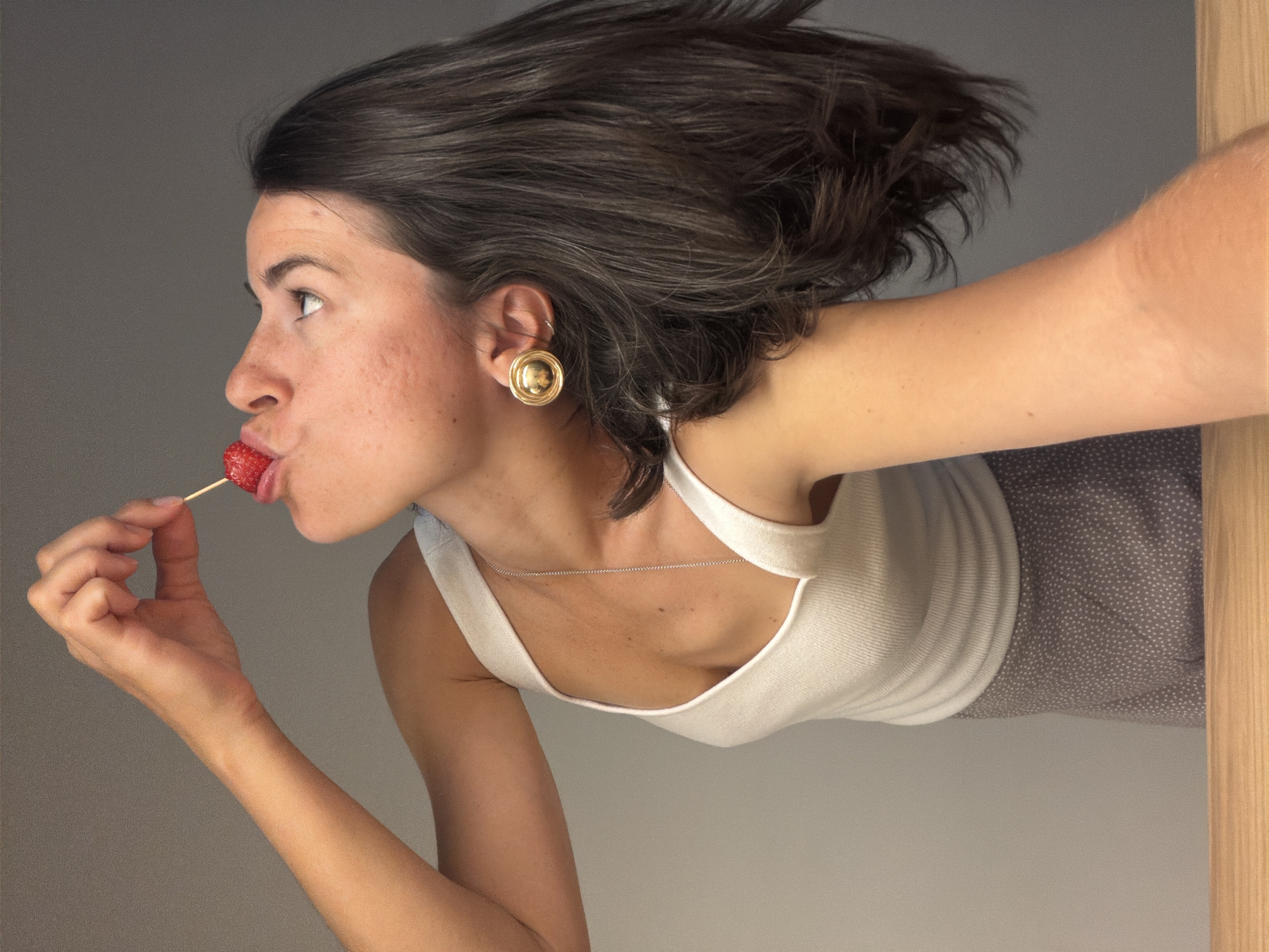 A woman with dark hair and fair skin is holding a strawberry on a toothpick to her lips. She is wearing a white tank top and gold earrings. The background is a plain gray wall. The bottom right of the image includes a wooden door frame and a gray polka dotted skirt. There is no indication of Budva, Montenegro.