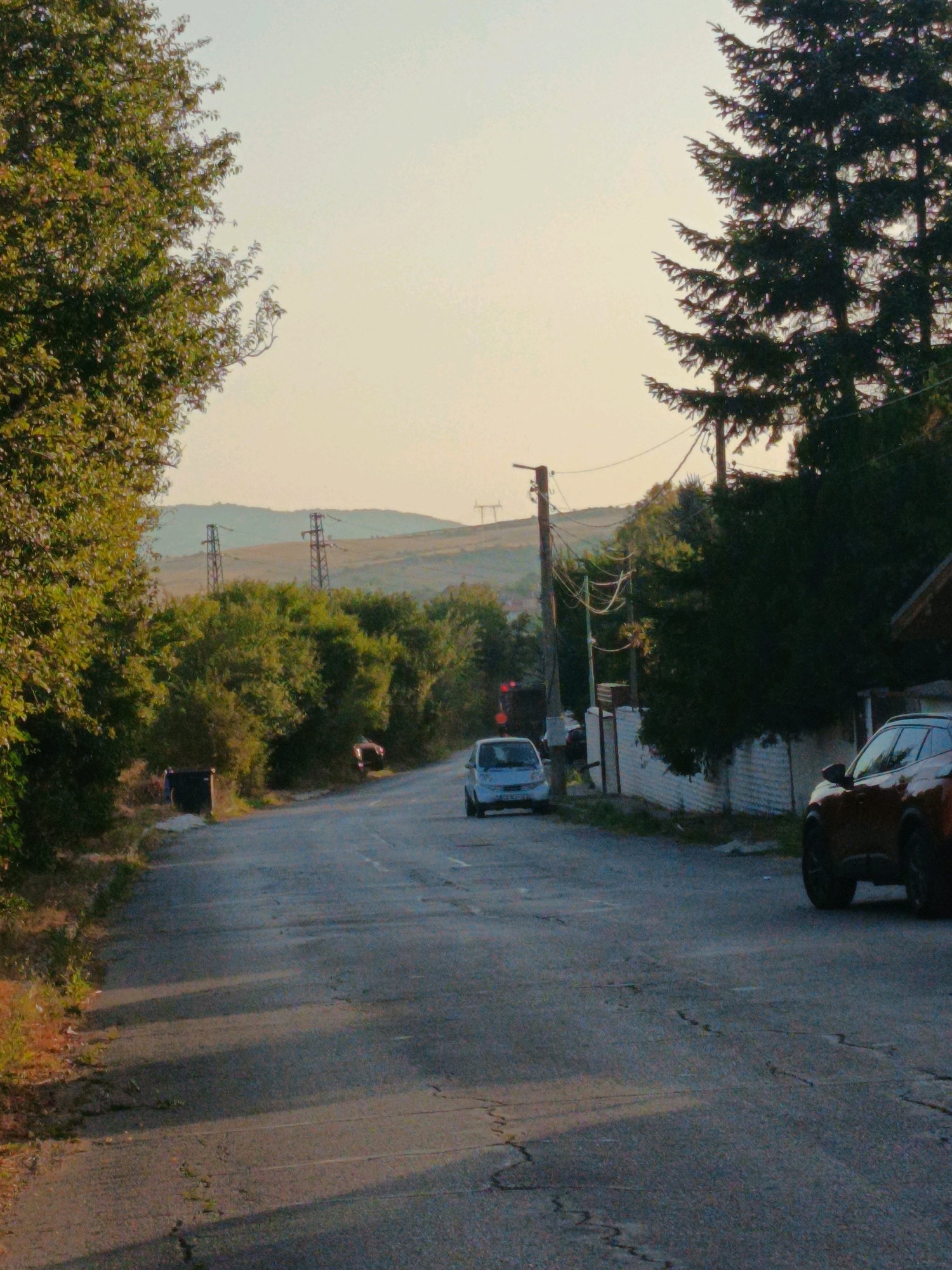 This is a medium shot, taken from a low angle, of a paved road in Kralev Dol, Bulgaria, with trees and hills in the background. The sun is low in the sky, casting long shadows across the road. On the left side of the road, there is a dark green tree with dense foliage, and a black trash receptacle. Further up the road, there are two cars parked on the right side, and a small white car on the left side. The road is cracked and weathered. In the background, there are hills with some vegetation and two power line towers. The sky is a hazy yellow-orange.