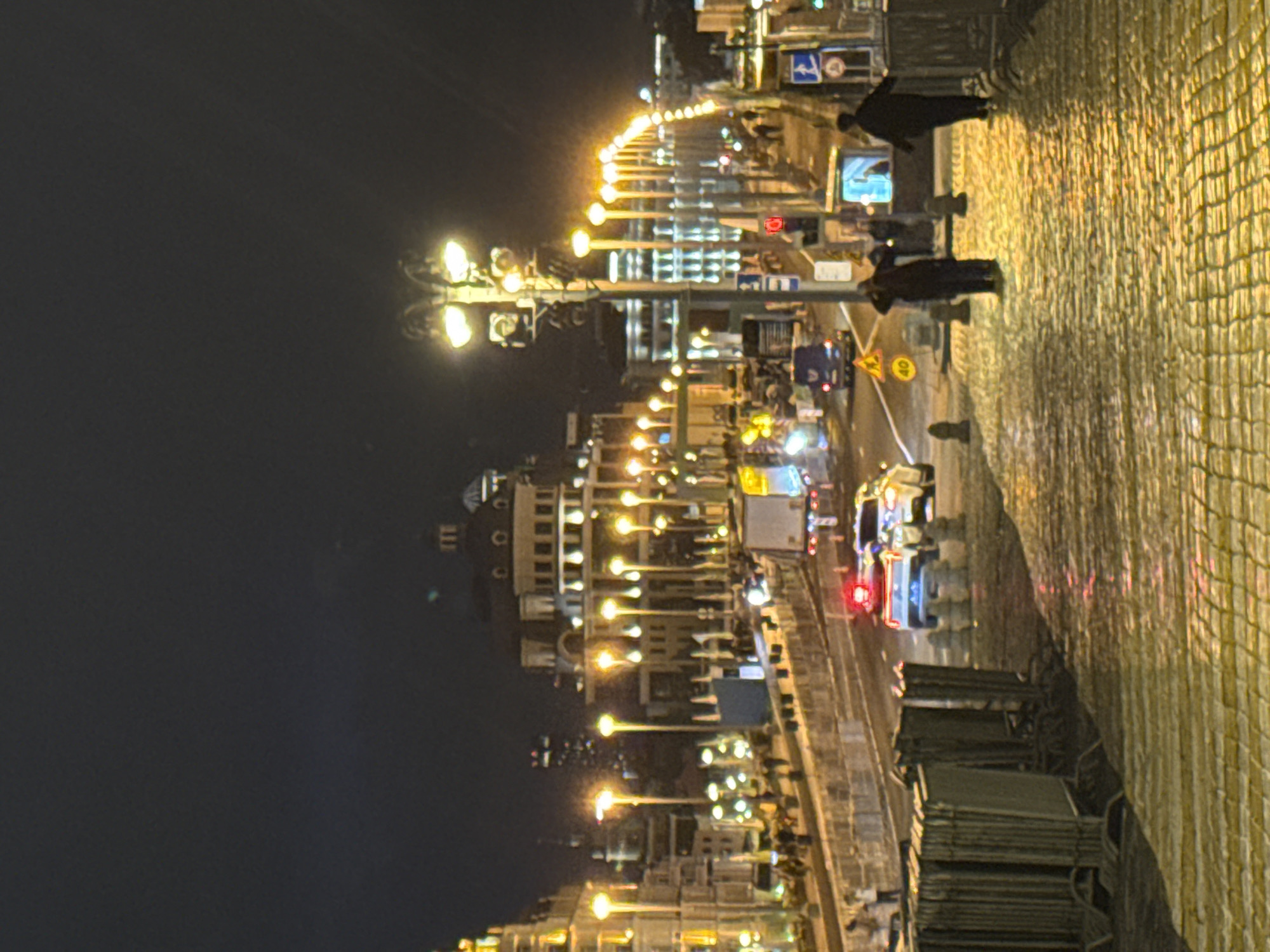 The image depicts a nighttime urban scene in Moscow, Russia. A cobblestone pedestrian walkway runs along the right side of the frame. A road stretches along the left side of the frame, lined with streetlights. Vehicles, including what appears to be a bus and cars, are visible on the road. People are walking along the walkway. Buildings illuminated with yellow lights are visible in the background, stretching into the distance. The sky is dark.