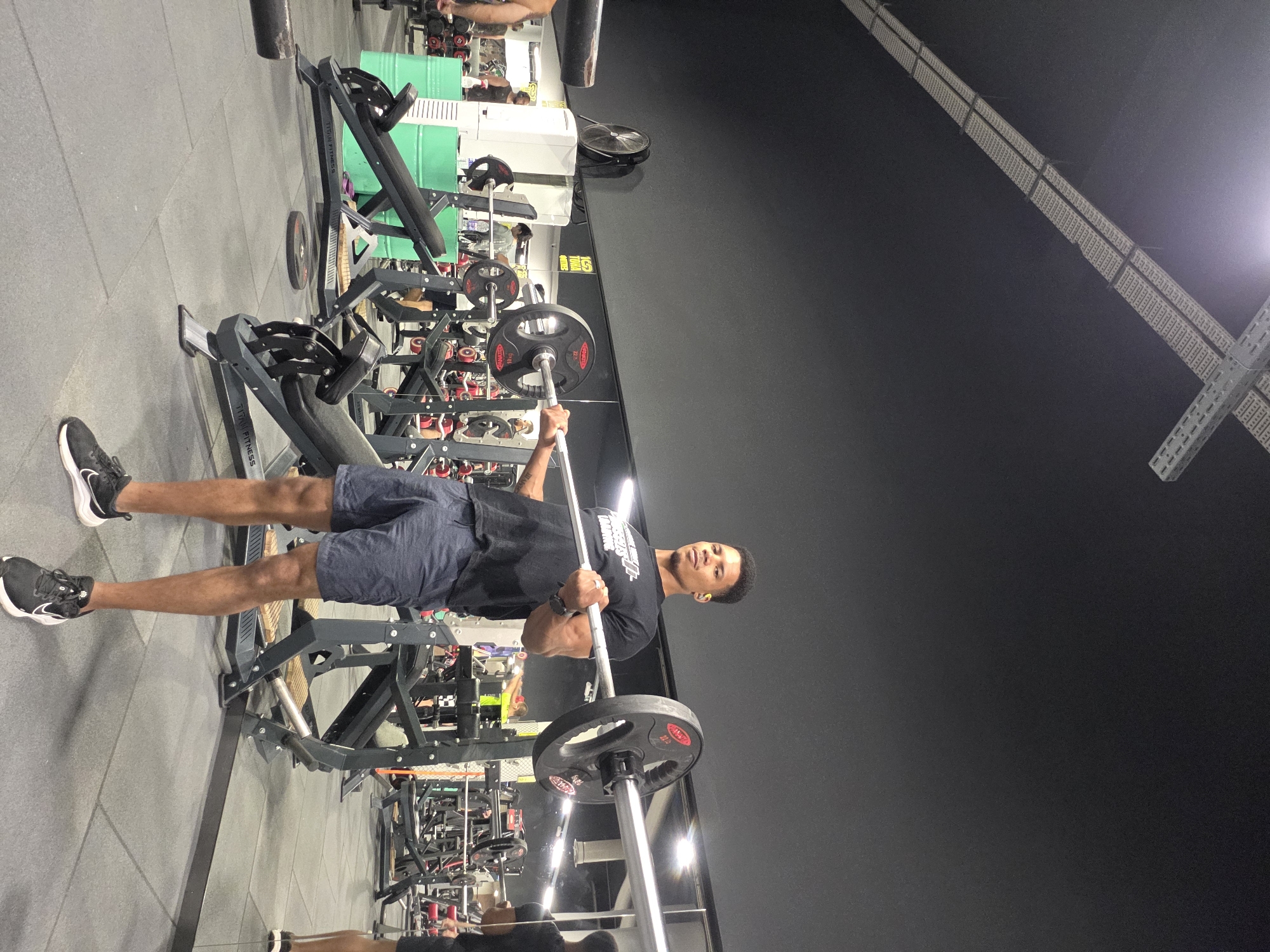 A young Black man is performing a barbell exercise in a gym in Varna, Bulgaria. He is standing and holding a barbell across his shoulders, with his arms bent and his hands gripping the bar. He is wearing a black t-shirt with white lettering and dark blue athletic shorts. His legs are slightly bent, and he is looking forward with a focused expression. The gym is well-lit, with a dark ceiling and grey tiled floor. Various weightlifting equipment, including benches, racks, and weights, are visible in the background, along with mirrors and a large industrial fan.