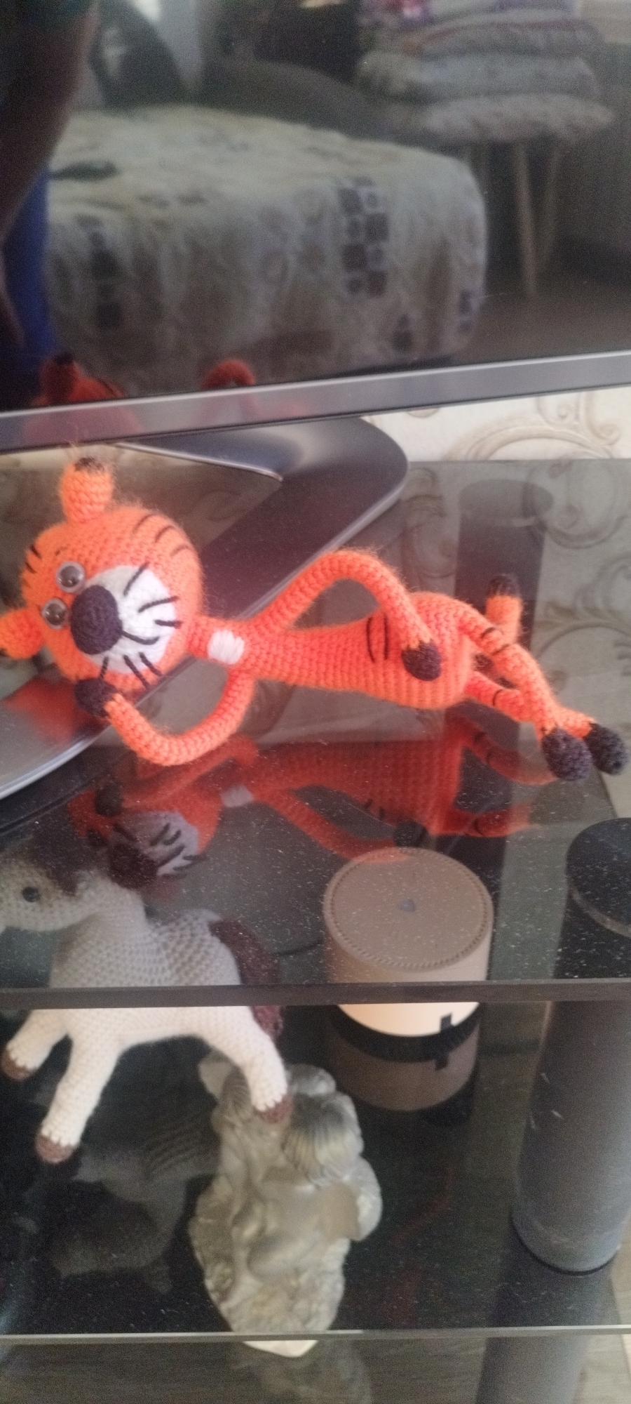 A handcrafted, orange-colored, knitted tiger toy with googly eyes and black stripes is lying on a reflective glass surface, likely a shelf. The tiger has a white patch on its chest and black paws. Its reflection is visible on the glass. Below the shelf, there are other objects, including a knitted white unicorn with brown hooves, a small grey statuette, and two cylindrical containers: one is tan with a black band, and the other is black. The shelf appears to be part of a piece of furniture, possibly a table or stand, with a television screen partially visible in the background. The blurred reflection in the upper portion of the image suggests the presence of other furniture and possibly a person taking the photo. There is nothing in the image to determine a precise location or confirm it being in Mokhsogollokh, Russia.