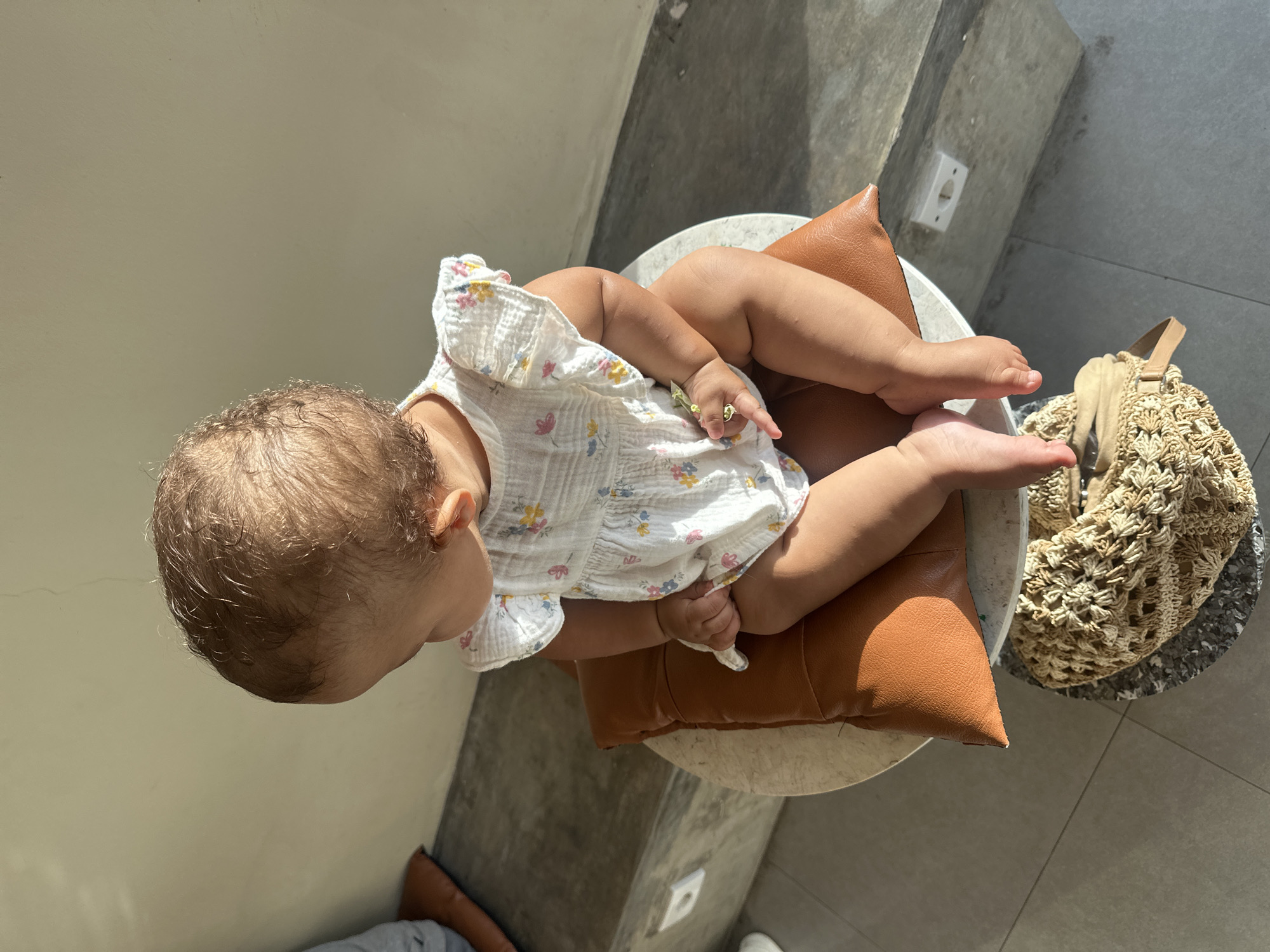 A baby in a white, floral-patterned dress sits on a brown pillow placed on a small, round, white table. The setting is outdoors, with a concrete wall and floor visible, suggesting a patio or terrace. A woven handbag rests on the floor near the table. The location is North Kuta, Indonesia.