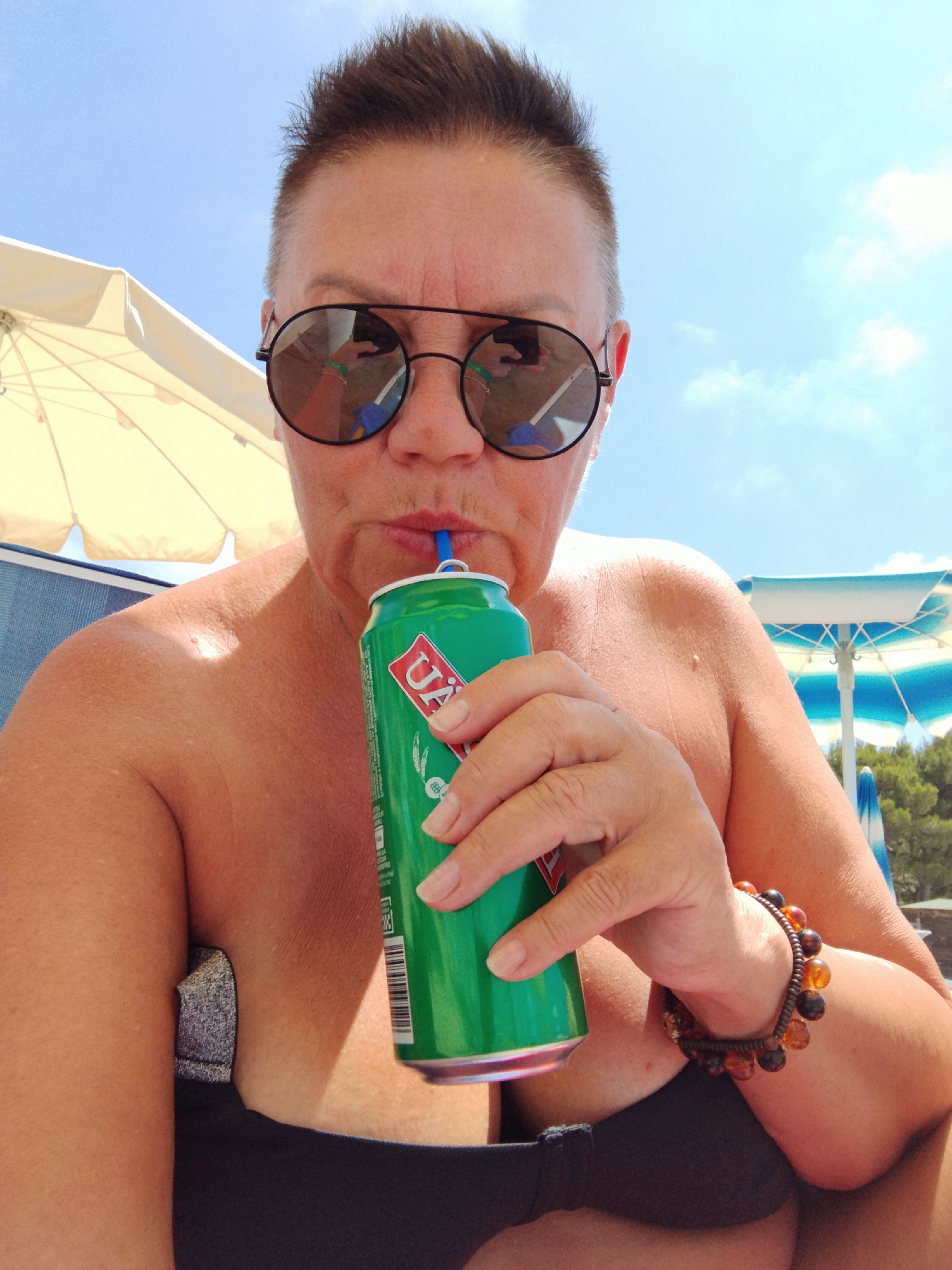 A close-up, low-angle, front-facing selfie captures a person with short, spiky hair and sunglasses, holding a green can with a blue straw to their mouth. The person is wearing a black bikini top and a beaded bracelet on their left wrist. The background suggests an outdoor setting with a beach umbrella and a clear blue sky with scattered clouds. The scene is likely Peschici, Italy, given the context provided.