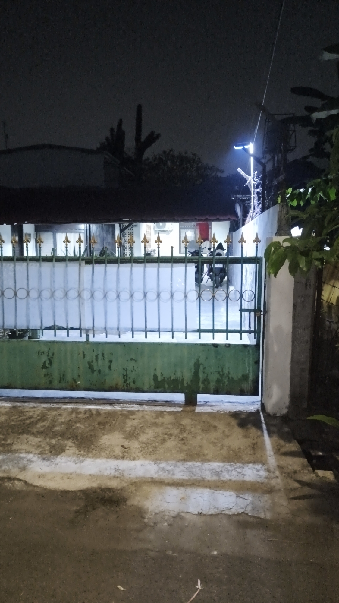 A residential gate in South Jakarta, Indonesia, is seen at night. The gate is made of green metal with a white panel in the upper section. Ornate gold finials top the vertical bars of the gate. Behind the gate, two motorcycles are parked. A street light illuminates the area. The ground in front of the gate appears to be wet, with white painted lines indicating a walkway. The background shows a dark sky and the silhouette of trees and buildings.