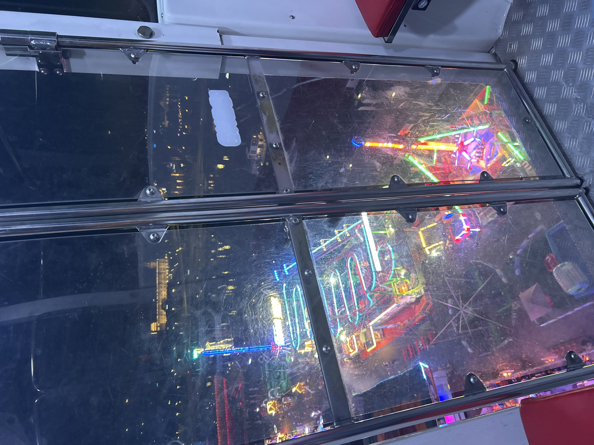 The image captures a nighttime view of an amusement park from within a transparent enclosure, likely a Ferris wheel cabin. The vibrant lights of various rides, including a towering illuminated structure with a blue glow and a spinning ride with colorful lights, illuminate the scene. In the distance, across a body of water, a large, illuminated building with a distinctive architectural style is visible, suggesting a prominent landmark in the city. The reflections and smudges on the glass create a slightly distorted but atmospheric perspective. The floor of the enclosure is a metallic diamond plate. The scene is set in Szczecin, Poland, likely during a festival or event.