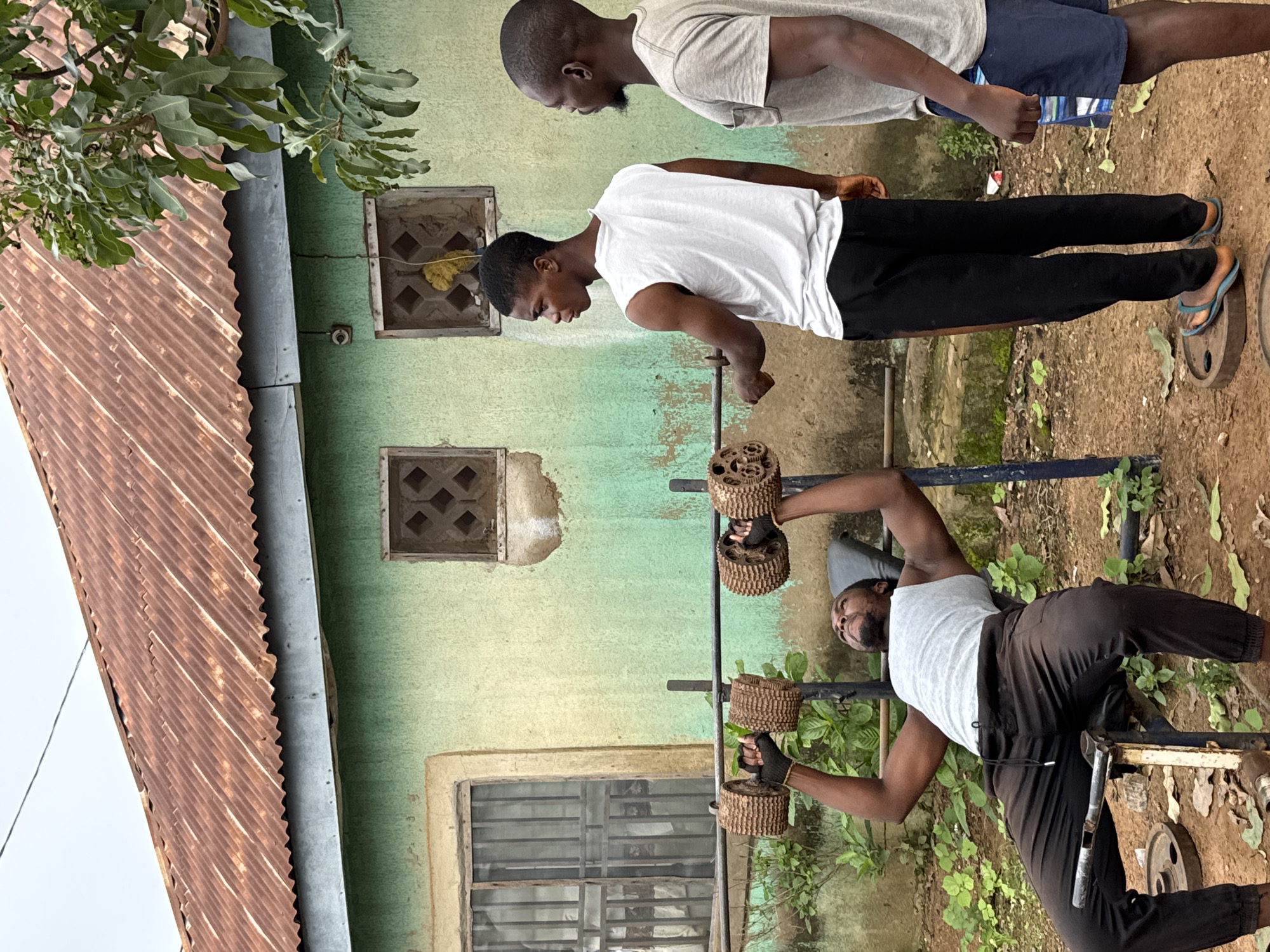 The image depicts an outdoor workout scene in Akwanga, Nigeria. Three young African men are present. One man is lying on a bench press, performing chest presses with a barbell equipped with makeshift, circular weights that appear to be made of repurposed materials. He is wearing a grey tank top and dark pants, with black gloves on his hands. A second man, wearing a white sleeveless shirt and black pants, stands to the side, observing the exercise. The third man, in a grey t-shirt and blue shorts, stands further to the right, also watching. The background features a weathered, green building with a rusty corrugated metal roof, two windows with decorative grilles, and some overgrown vegetation. The ground is composed of reddish-brown earth. The overall scene suggests a simple, informal gym setup in a residential area.