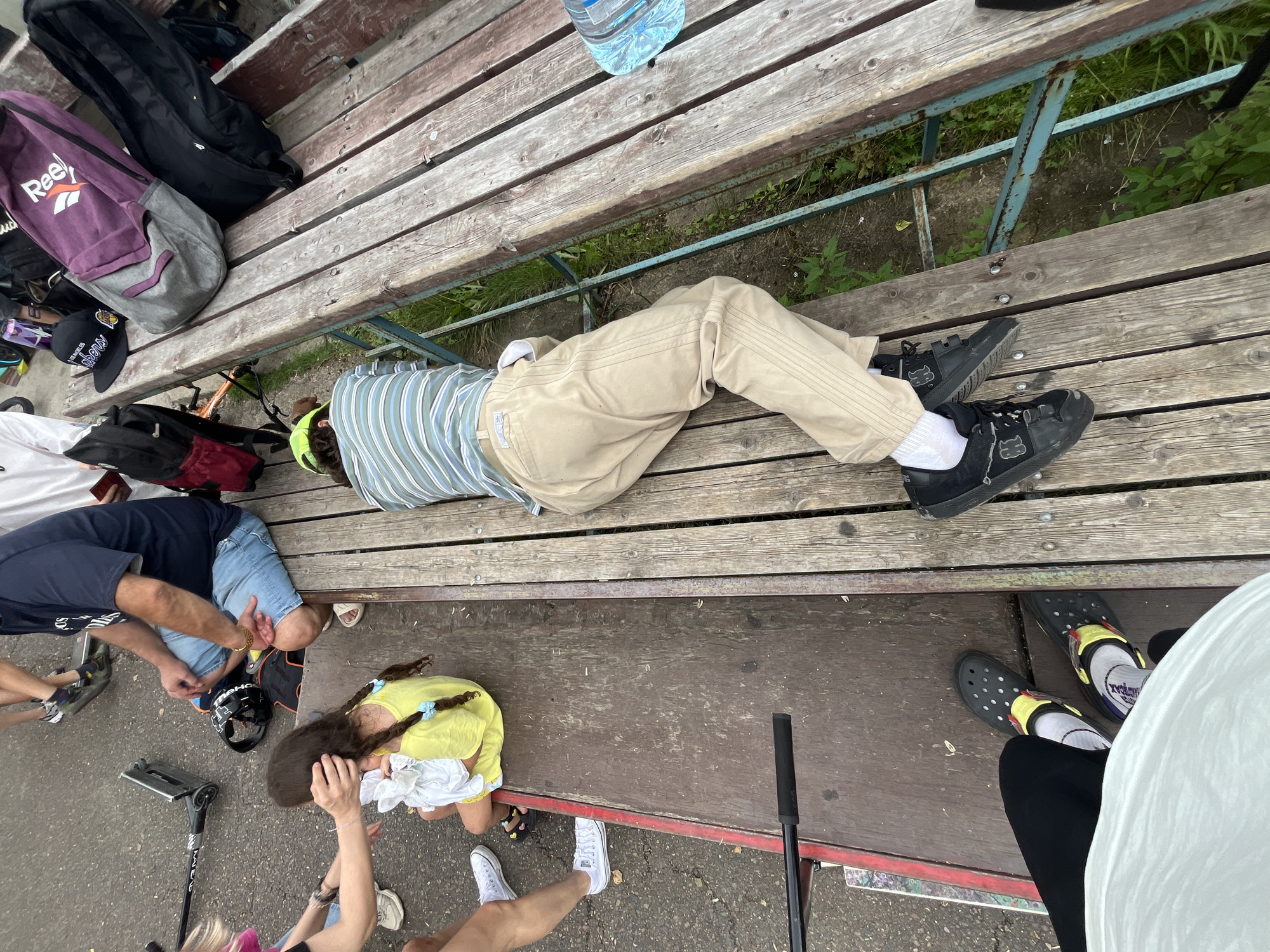 In Divnogorsk, Russia, a high-angle outdoor shot captures a scene on a weathered wooden bench. A person is lying on their stomach, facing away from the camera, with their head resting on the bench. They are wearing a striped t-shirt, khaki pants, white socks, and black sneakers. To the left of the bench, a young girl in a yellow dress sits with her legs crossed, holding a white bundle. Nearby, a scooter is parked on the ground. Several bags, including a purple Reebok bag and black backpacks, are placed on the bench and ground. The environment appears to be a park or public space, with grass and dirt visible around the bench.