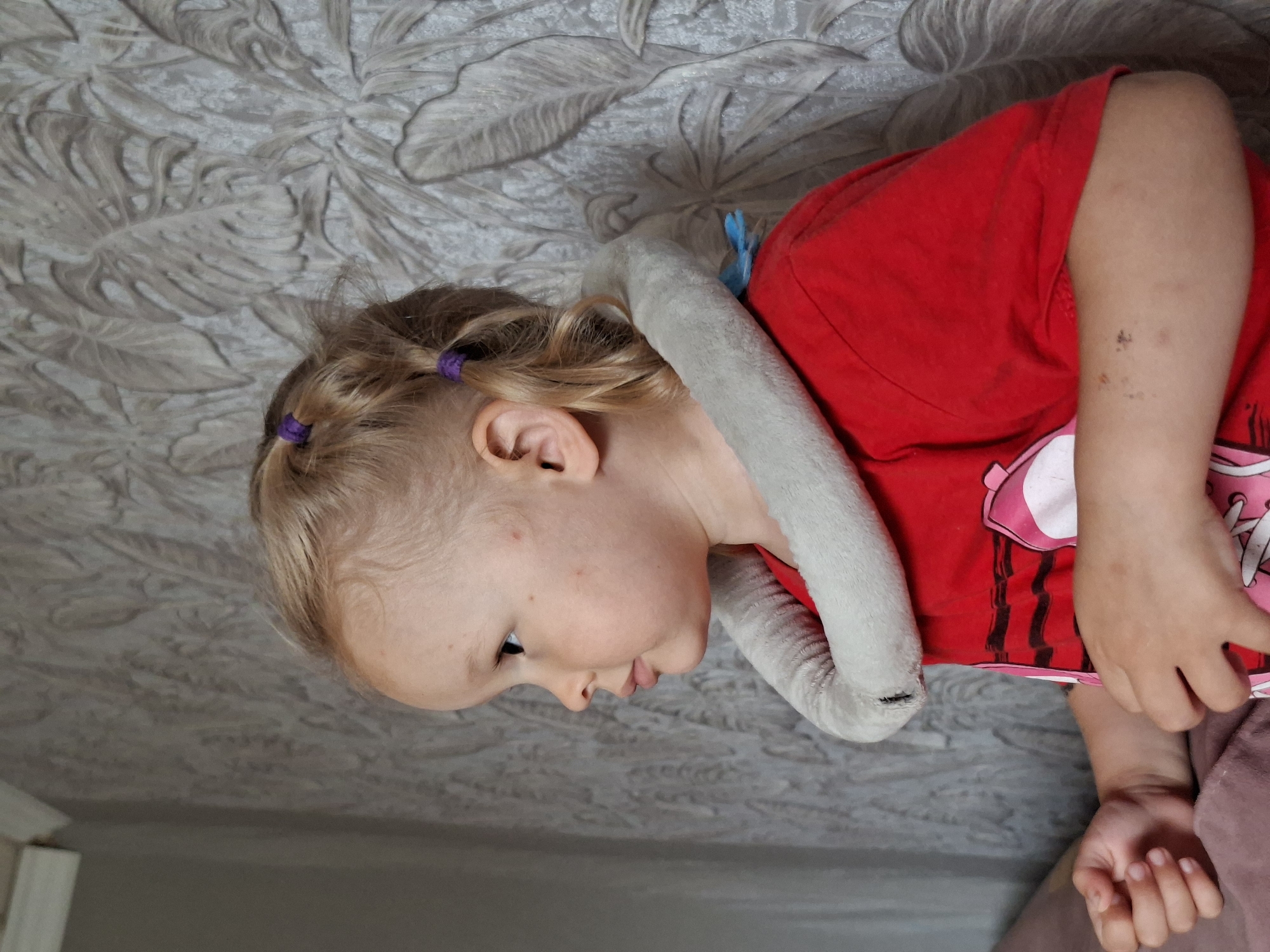 A medium shot shows a young child with blonde hair styled in two pigtails with purple hair ties, facing left in profile. The child is wearing a red t-shirt and a grey, plush neck pillow. The background consists of a wall with a textured, grey floral wallpaper pattern. The lighting suggests an indoor setting. The image is a still photograph.