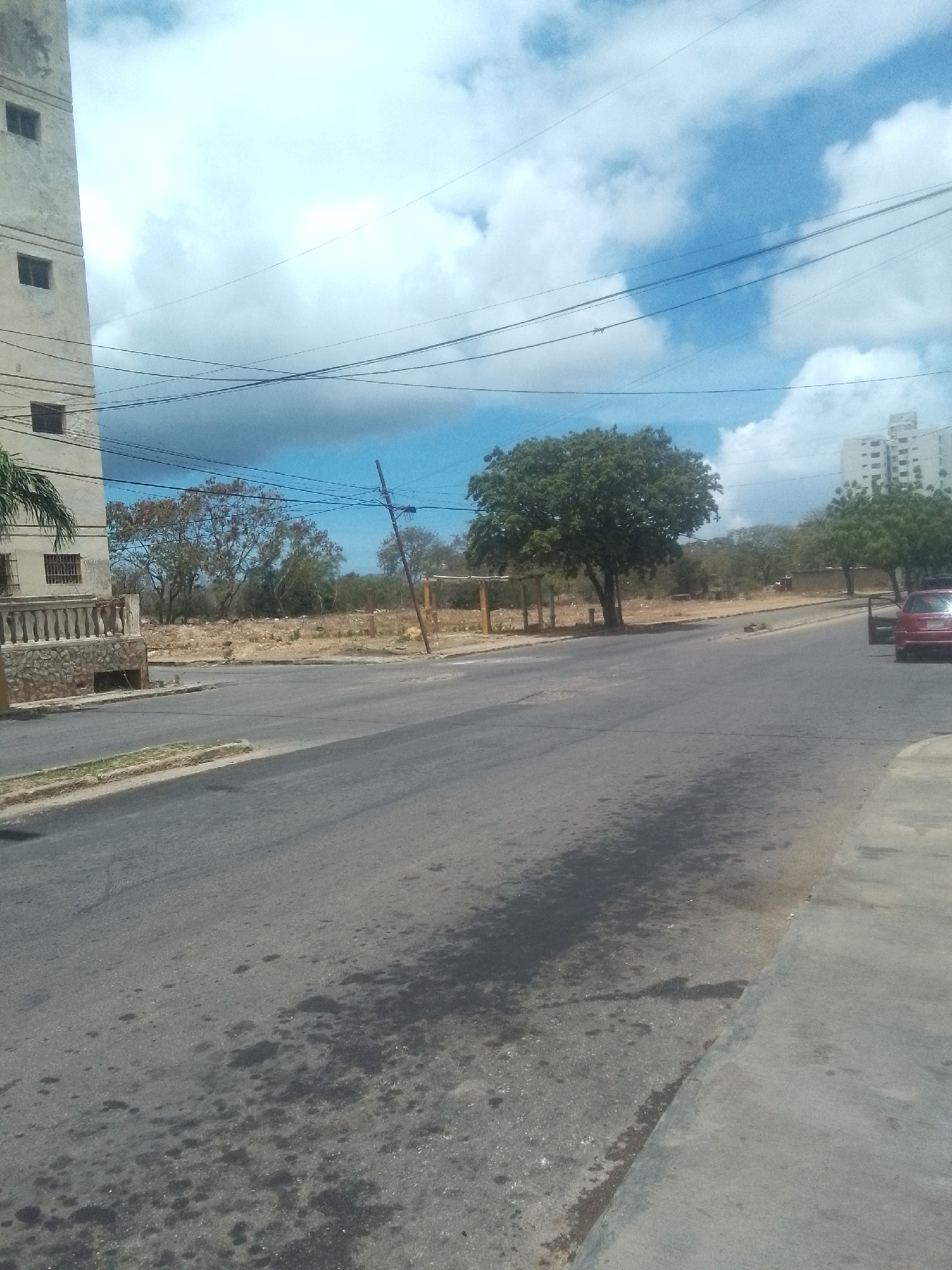 The image shows a street scene in Juan Griego, Venezuela. A multi-story building stands on the left, with another in the distance to the right. A red car is parked on the right side of the street. Trees line the street and an empty lot is situated in the middle ground, behind a rustic fence. Overhead wires crisscross the sky, which is partly cloudy with patches of blue. The asphalt road appears damp, showing darker areas and various splatter stains. The sidewalk is visible on the right edge of the frame.