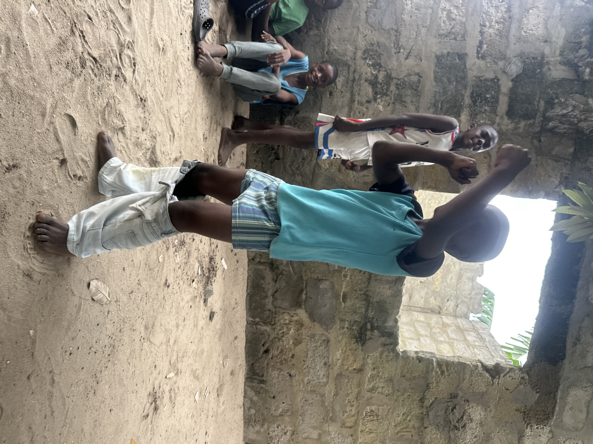 In an outdoor setting with sandy ground and a rough stone wall, three young boys are visible. One boy in the foreground, on the left, is wearing light-colored pants and a turquoise t-shirt. He is facing away from the camera and appears to be in the process of standing up, with his left leg extended and his right leg bent. His arms are raised, and his right fist is clenched.
Behind him, another boy is seated on the sand, wearing a blue t-shirt and grey pants. He is smiling and looking towards the camera, with his hands resting on his knees. To his right, a third boy is standing, wearing a white tank top with red and blue accents and striped shorts. He is looking towards the boy in the foreground and has his hands raised in a gesture that resembles a fist bump.
The scene appears to be an informal gathering, possibly in a village or rural area given the sandy ground and rustic stone wall. The lighting suggests it is daytime, with diffused light indicating it might be overcast or late afternoon. The overall mood seems lighthearted and playful, with the smiles and gestures of the boys conveying a sense of camaraderie. The presence of leaves on the ground and greenery visible through an opening in the wall suggests a natural, possibly tropical, environment. No visible text is present in the image.
