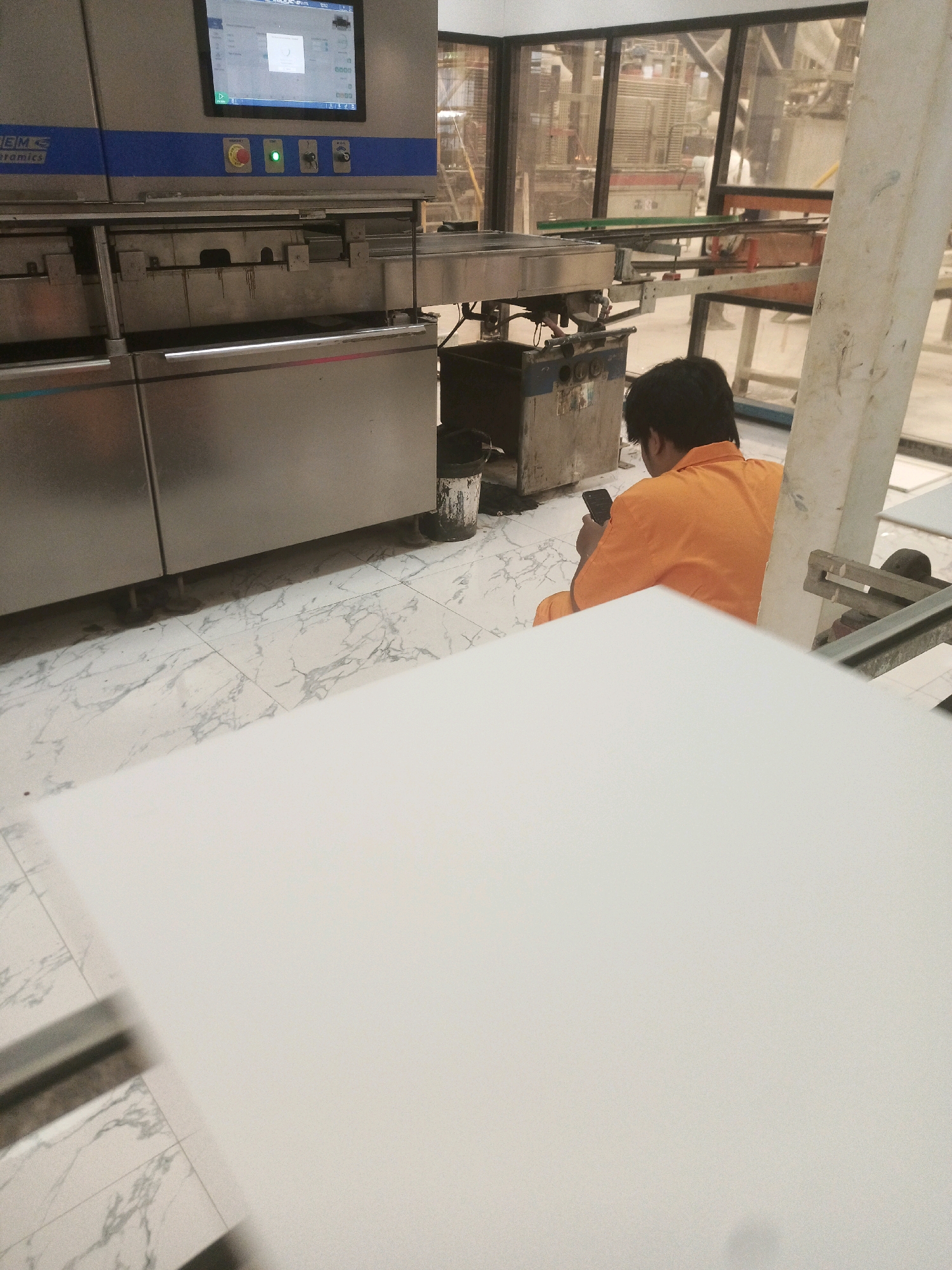 In Lebaniwaras, Indonesia, a man in an orange jumpsuit sits on the floor of what appears to be a factory or industrial setting. He is looking at a mobile phone in his hand. In the background, large industrial machinery with a digital display screen is visible, along with various metal components and structures. The floor is tiled with a marble-like pattern. A waste bin is positioned near the machinery.