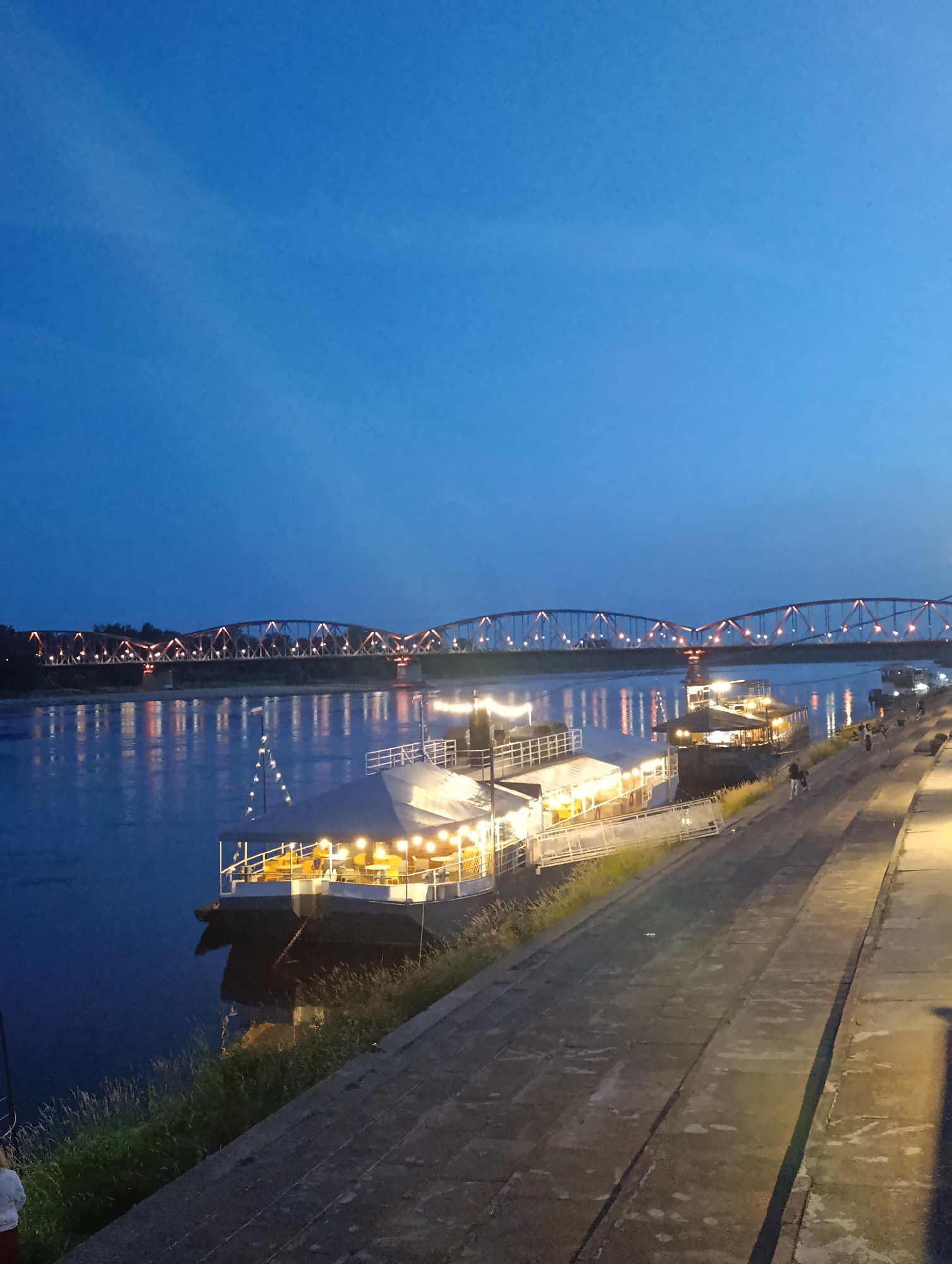 A long shot captures a twilight scene along the Vistula River in Toruń, Poland. A large, illuminated passenger boat is docked along the concrete embankment, its lights reflecting on the calm water. Further in the distance, a wide, truss bridge spans the river, its structure outlined by numerous amber lights. The sky is a deep twilight blue with a hint of clouds. Several figures are visible on the embankment, some walking, others standing near the boat. The overall atmosphere is serene and picturesque, with the man-made structures illuminated against the natural backdrop of the river and sky.