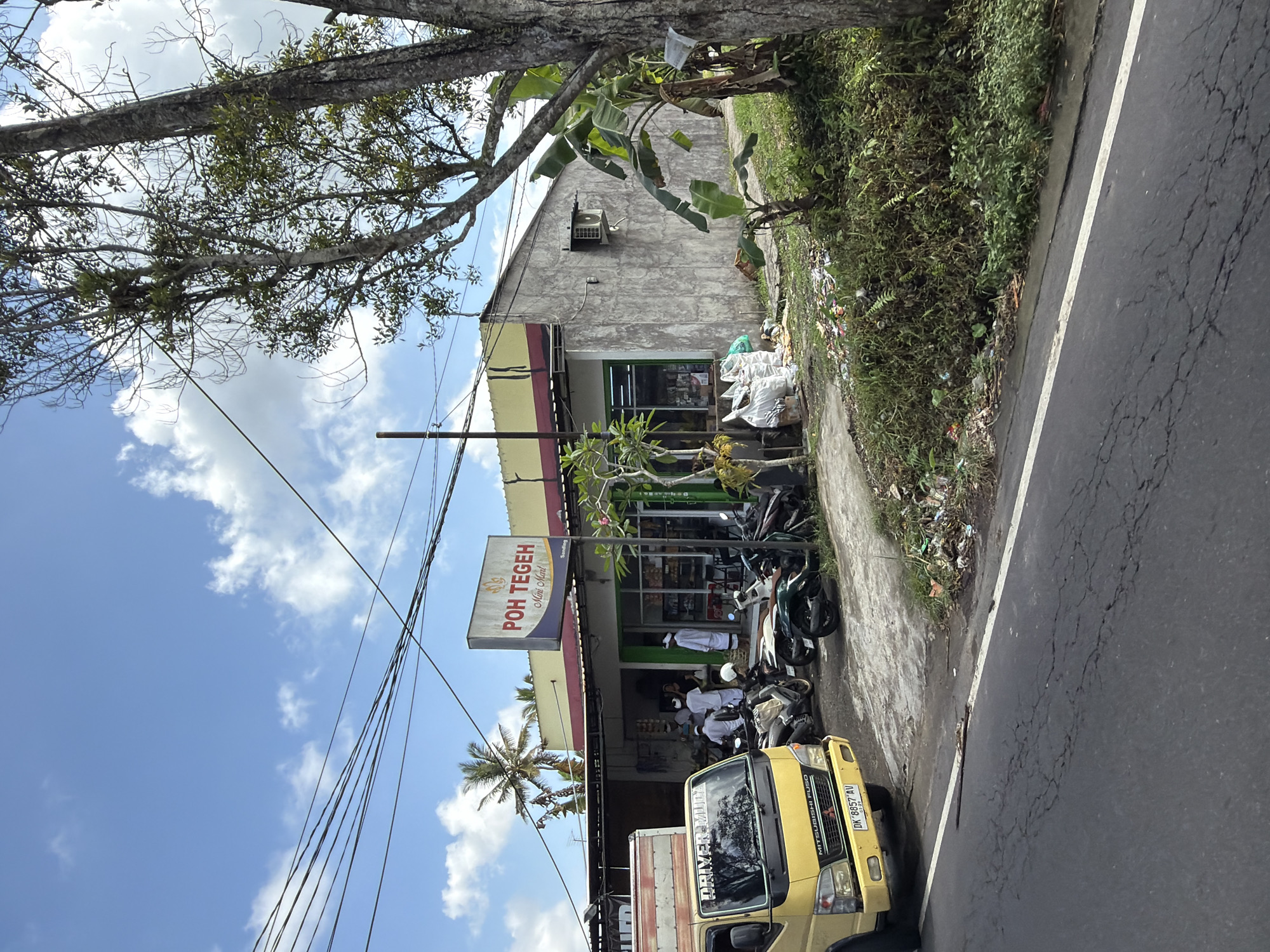 This is a medium shot, outdoor, eye-level, daytime photograph of a small convenience store in Tampaksiring, Indonesia. The store is a single-story building with a yellow awning over the entrance and a gray concrete exterior. A sign above the entrance reads "POH TEGEH Mini Mart." Several people, some wearing traditional white clothing, are standing near the entrance of the store. A yellow Mitsubishi Fuso truck is parked to the left of the store, with "DRIVER Muly" written on the windshield. Several motorcycles are parked in front of the store. The ground in front of the store is a mix of pavement and overgrown vegetation, with some litter visible. A large tree with lush green leaves is on the right side of the image, and power lines are visible in the sky above. The sky is blue with scattered white clouds.