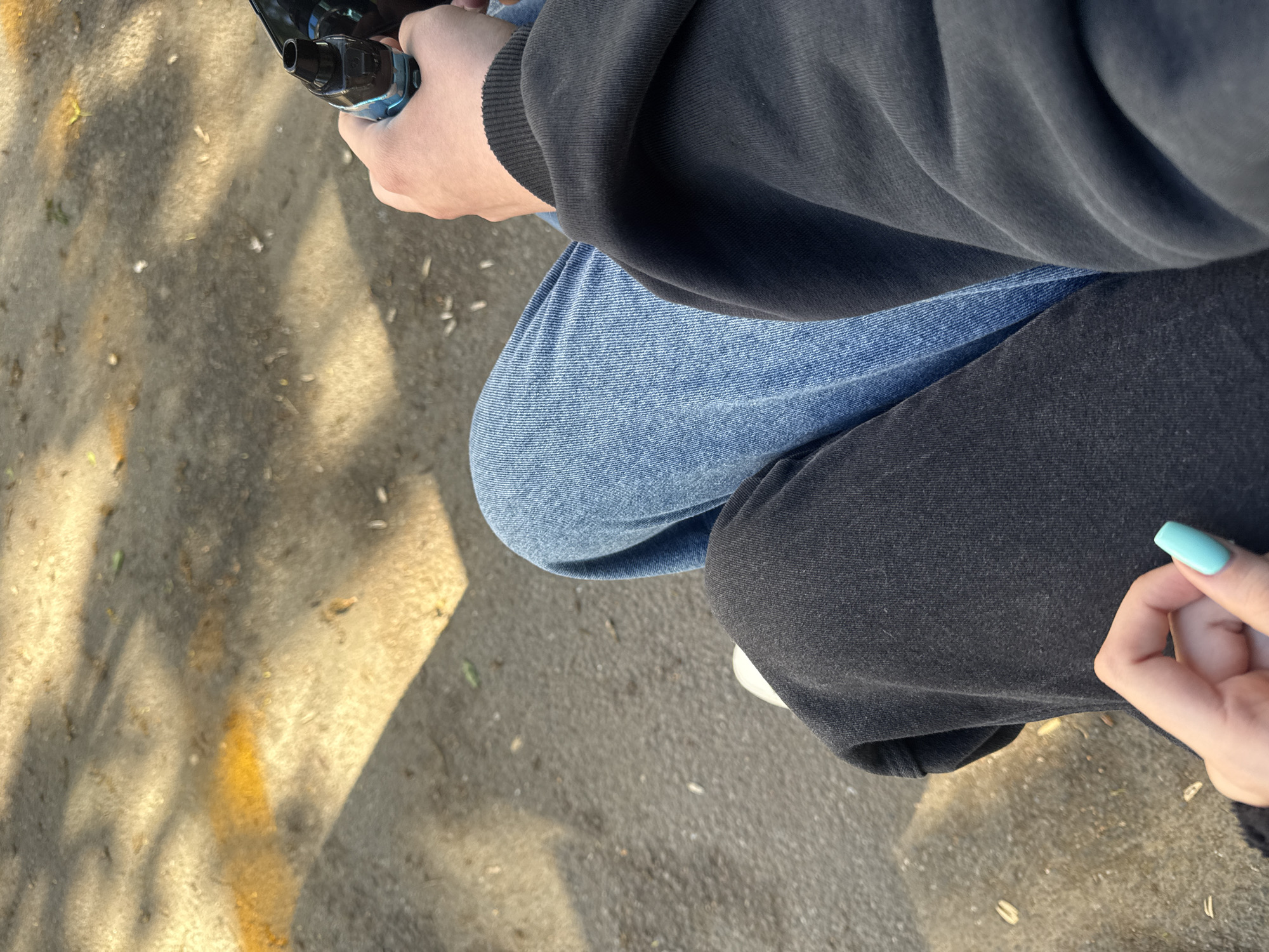An overhead shot depicts a person seated outdoors, possibly on the ground in Severnaya Samarka, Russia. The individual wears a dark-colored hooded sweatshirt and blue denim jeans on one leg, while the other leg is clad in dark-colored pants. The person holds a vaping device in their left hand. The ground is a light-colored surface. A glimpse of a hand with a light blue nail is visible near the person's right leg.