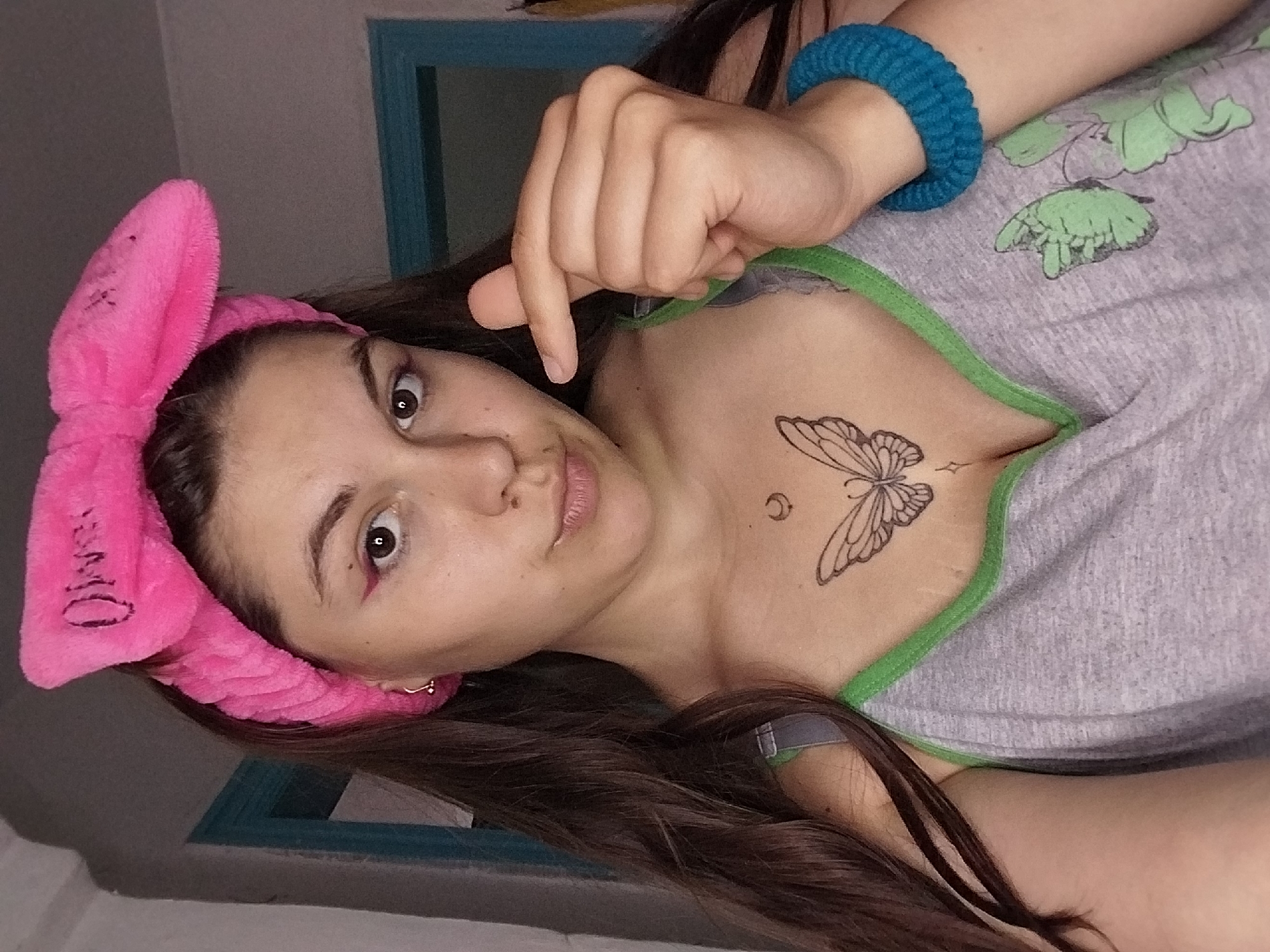 This is a vertically oriented, close-up, selfie-style photograph of a young woman. She is lying down, with her head tilted and turned towards the camera. The woman has long, dark brown hair, and she is wearing a bright pink, fuzzy headband with a large bow on top. The headband has black lettering visible on the bow, which appears to be "OME." She is also wearing a grey t-shirt with green trim around the neckline. A blue, knitted bracelet is visible on her right wrist. She has a tattoo of a butterfly on her chest, just below her collarbone, and a small crescent moon tattoo to the left of the butterfly. Her makeup includes dark eyeliner and pink lipstick. Her eyes are wide open and she is looking directly at the camera with a neutral expression. Her right hand is visible in the upper right portion of the frame, with her index finger pointing downwards. The background is out of focus, but appears to be an indoor setting with a grey wall and a window frame with blue trim. The lighting suggests it is daytime, but the indoor setting provides no clear indication of weather or specific location. The overall mood of the image is casual and personal.