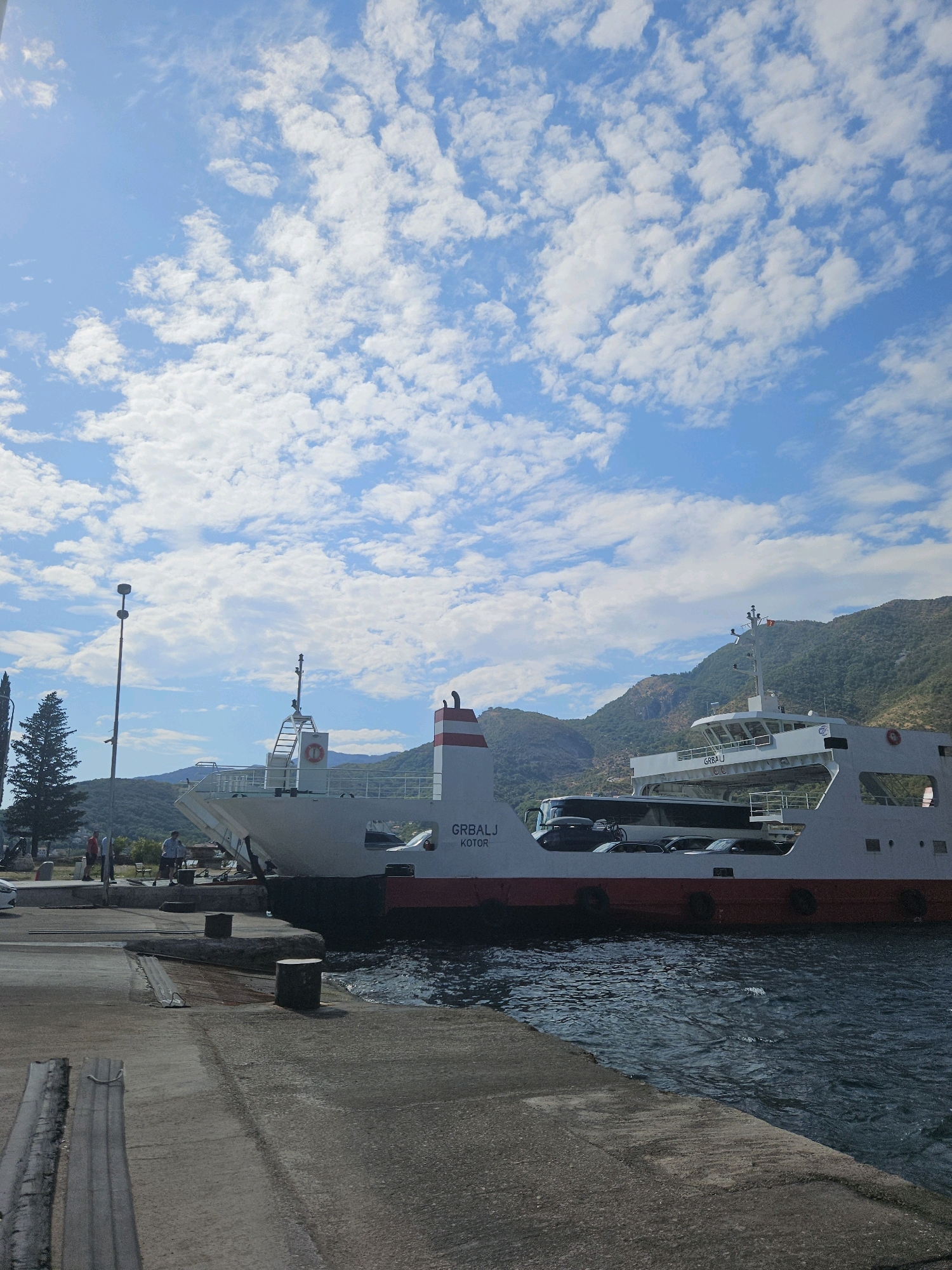A ferry named "GRBALJ KOTOR" is docked at a port under a partly cloudy blue sky. The ferry, loaded with cars and a bus, is white with a red stripe along its waterline. Mountains form the backdrop. On the shore, a concrete pier extends into the water. Several people are visible on the pier, near a lamppost and some trees. The water is choppy.