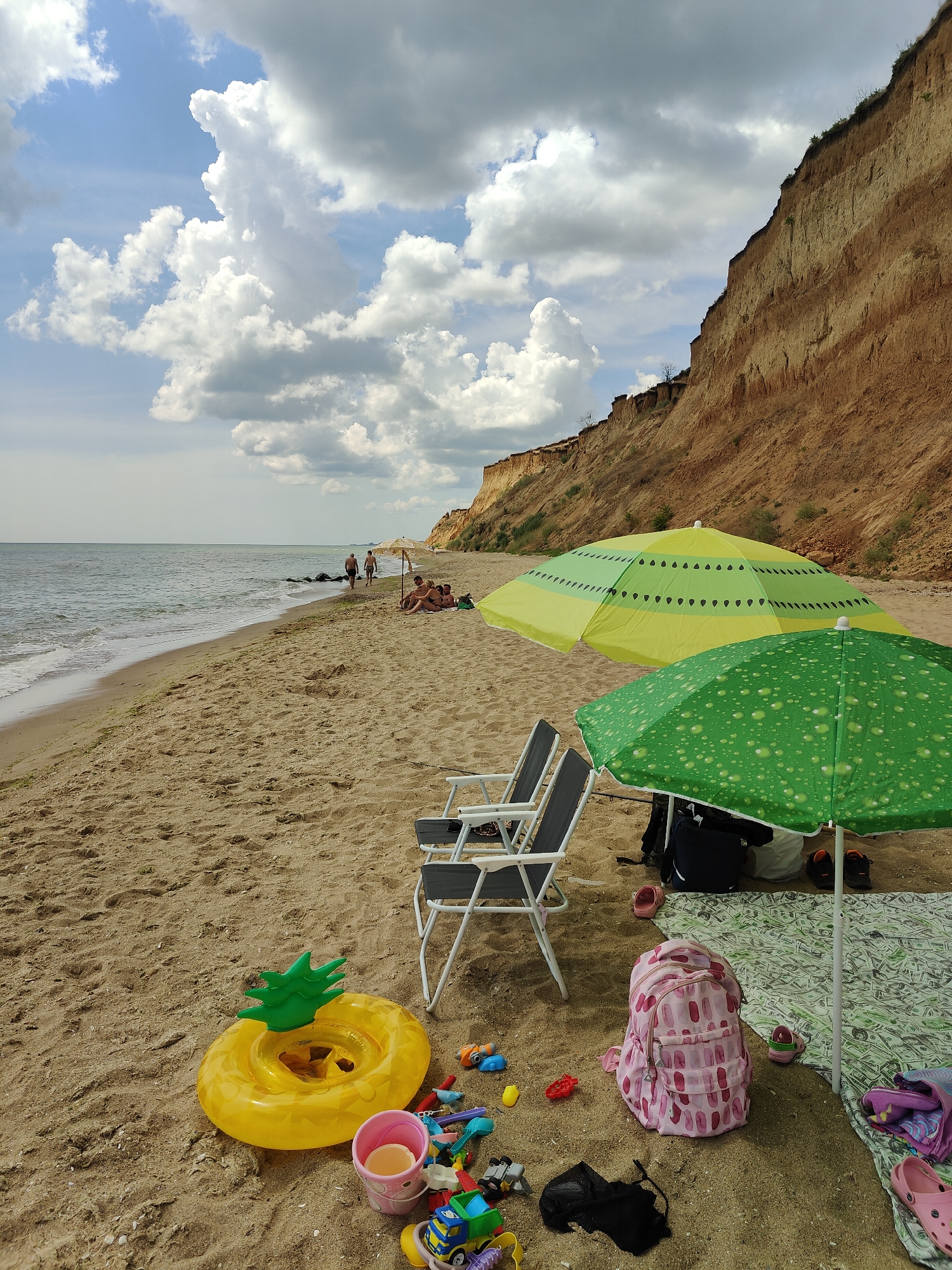This is a daytime, eye-level, full-shot photograph taken on the sandy beach of Sanzhiika, Ukraine. In the foreground, a yellow inflatable pineapple-shaped ring, a pink bucket filled with sand, and various children's toys are scattered on the sand. Two empty beach chairs are positioned to the right of these items, with a large green umbrella with a bubble pattern and a yellow umbrella with a watermelon pattern set up behind them. A patterned blanket is spread out under the green umbrella, and a pink and white patterned backpack rests on top of it, alongside some footwear and bags. Further back on the beach, a group of people are seated on the sand under a natural-looking umbrella, and two individuals are walking along the shoreline. The sea is visible to the left, with gentle waves reaching the shore. The background is dominated by a steep, sandy cliff face with visible layers of sediment, a common geological feature of the Sanzhiika coastline. The sky is partly cloudy, with dramatic white and gray clouds against a blue backdrop.