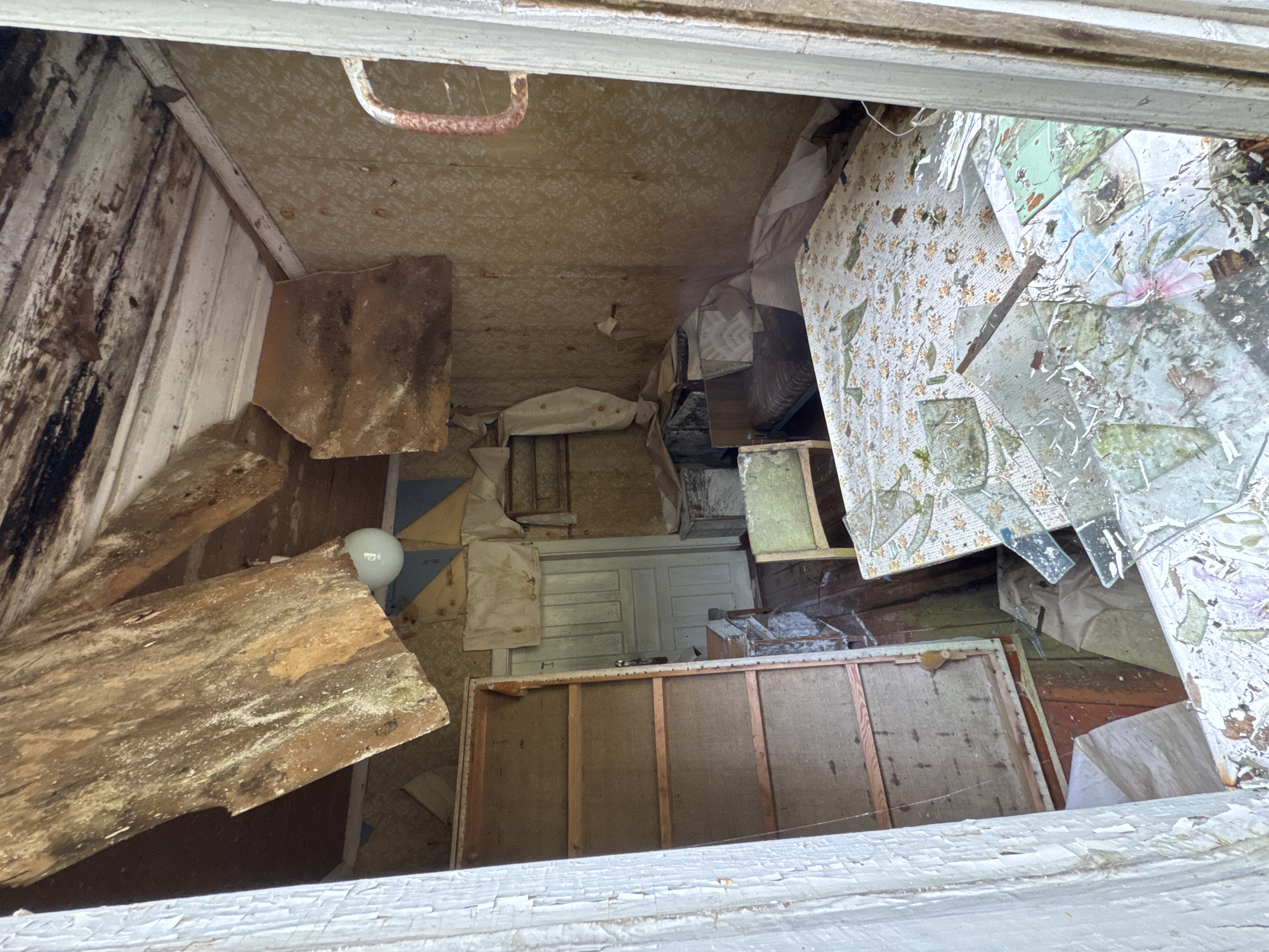 This is a full-frame, eye-level, interior shot of a derelict room in a house in Ikhala, Russia, taken in daylight. The room appears to be a bedroom or living area, with signs of severe decay and abandonment.
The ceiling is heavily damaged, with wooden planks and insulation hanging down, exposing the rafters and interior structure. Some of the ceiling material is water-damaged and discolored. A bare light bulb hangs precariously from a wire.
The walls are covered in faded, patterned wallpaper, some of which is peeling away. There are also diamond-shaped patterns of what appears to be fabric or paper applied to the upper portions of the walls.
The room contains several pieces of furniture in a state of disrepair. To the left, there is a tall, open shelving unit with wooden planks. In the center, there is a small, light green chair. In the background, there is a bed with a patterned bedspread. A table with a similar floral patterned cover is in the foreground. The surface of this table is covered in shattered glass, debris, and what appears to be shattered ceramic or porcelain.
A white-paneled door is visible in the background, slightly ajar. To the right of the door, there is a metal pipe or rod extending from the wall.
The floor is covered with debris, fallen ceiling material, and a large amount of shattered glass, particularly in the foreground. The overall impression is one of significant neglect and deterioration.