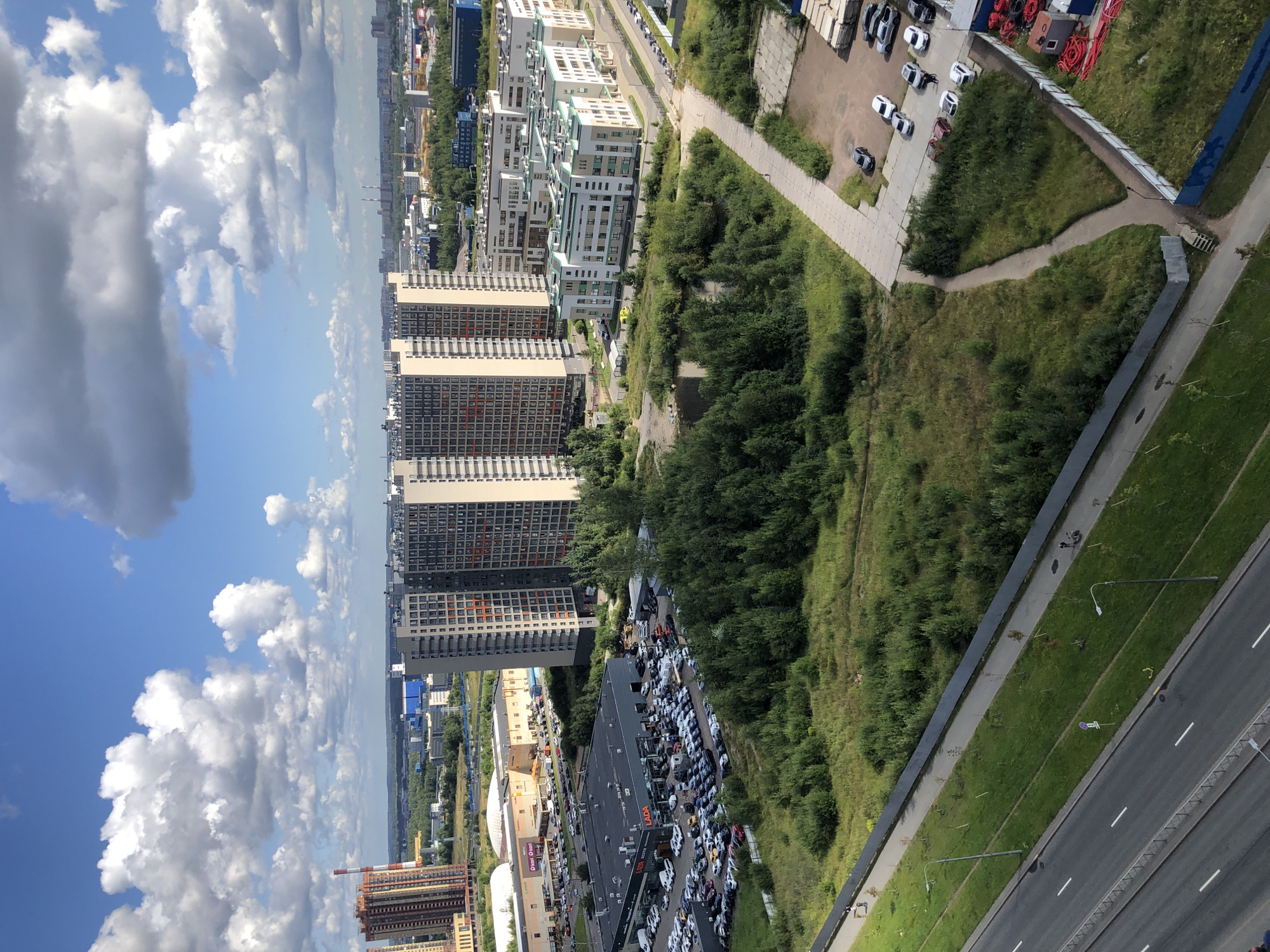 An aerial, medium shot captures a sprawling urban landscape under a bright, partly cloudy sky. The foreground features a multi-lane highway, with cars visible on the road and adjacent green, grassy verges. Beyond the highway, a large, undeveloped area is dominated by dense, green trees, with a small body of water. Several modern, multi-story residential buildings are situated throughout the scene, some with distinctive orange window accents. A large retail complex with a large parking lot filled with cars is visible to the left of the image, and a Lada car dealership is clearly marked. In the background, more buildings and industrial structures stretch towards the horizon, interspersed with trees and other vegetation. The overall impression is one of ongoing urban development and a mix of residential, commercial, and natural elements.