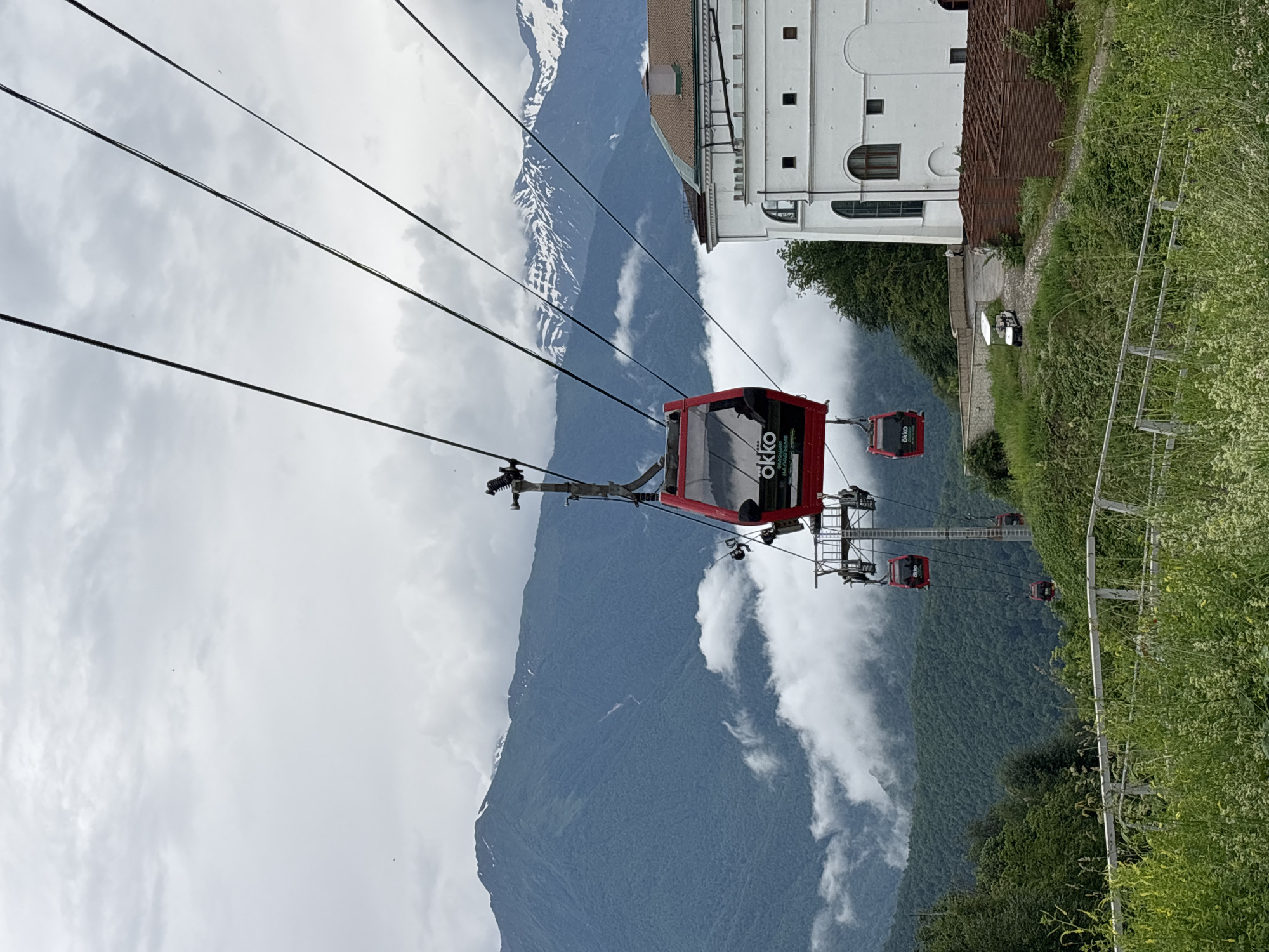 A red cable car cabin, branded "Okko," is suspended mid-air against a backdrop of misty, green mountains and a cloudy sky. The cable car is part of a system that travels along thick, dark cables, several of which are visible crisscrossing the frame. In the foreground, lush green foliage and wildflowers grow along a white railing. To the right, a modern white building with arched windows and a brown-tiled roof stands prominently. A small white golf cart is parked on a paved path leading to the building, with two individuals visible near it. Further down the mountain, other cable car cabins are visible, descending into the mist. The scene captures a view of a mountainous resort area in Estosadok, Russia.