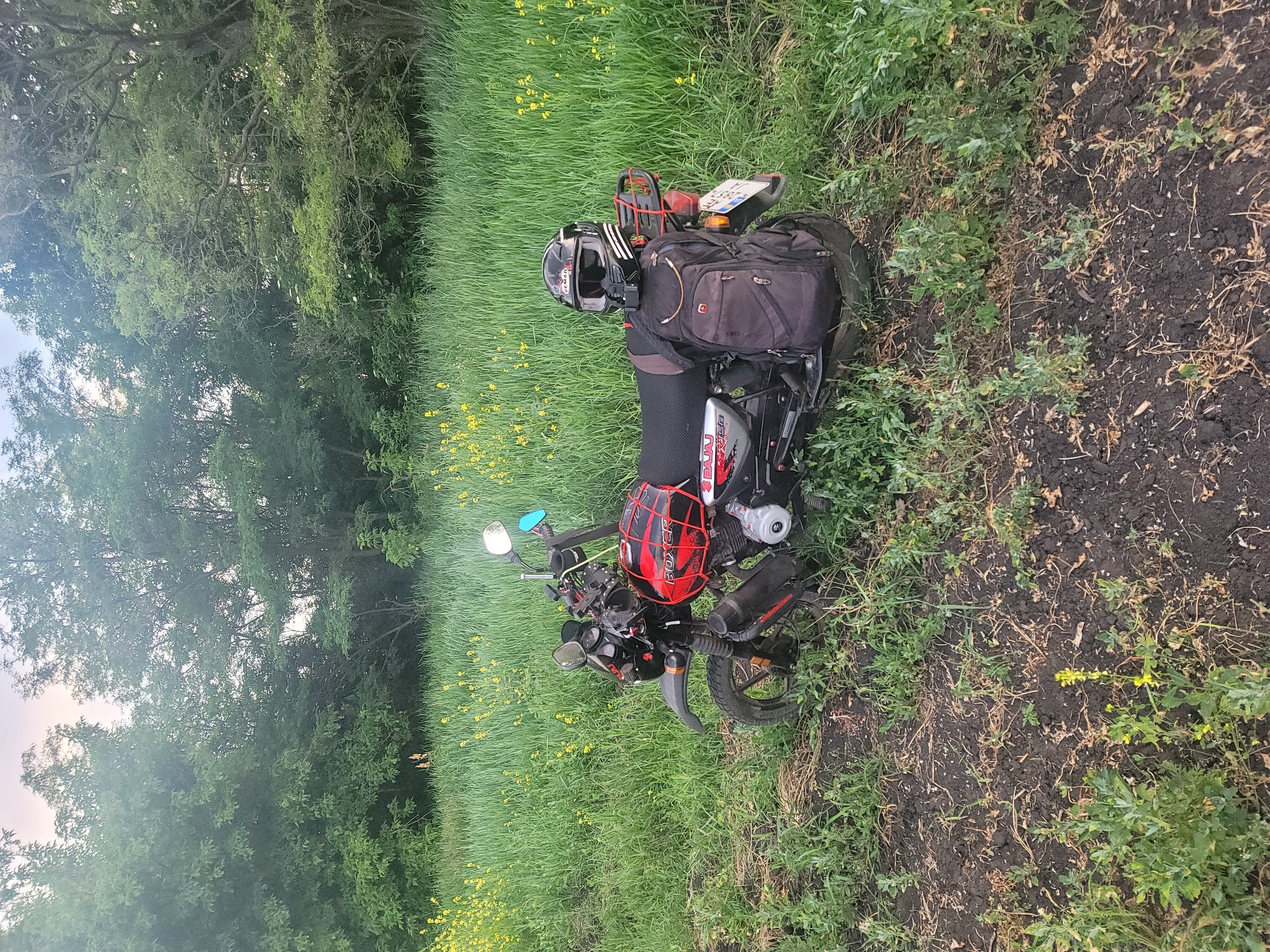 An overhead shot shows a red and black Bajaj Boxer motorcycle parked on a dirt path at the edge of a grassy field with yellow wildflowers. A black helmet rests on the motorcycle's rear seat, which also supports a black backpack. Trees line the background of the scene.