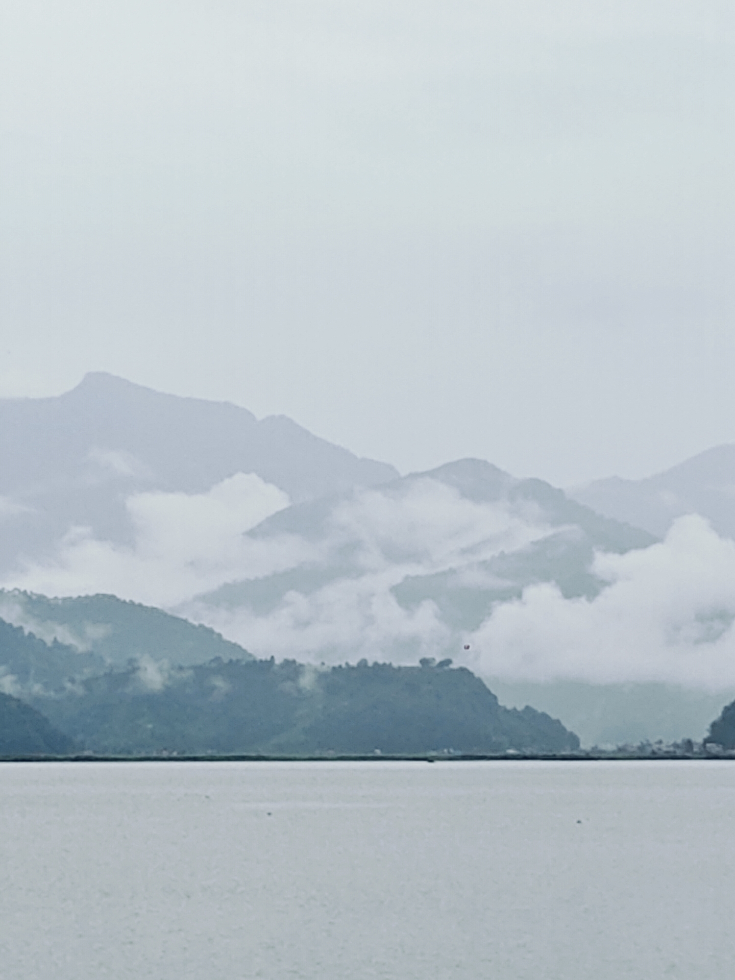 A panoramic view of the Annapurna mountain range from Pokhara, Nepal, captures the serene beauty of a hazy day. The foreground is dominated by the calm, reflective waters of Phewa Lake, its surface slightly rippled, hinting at a gentle breeze.
In the mid-ground, lush green hills rise, their slopes covered in dense vegetation. Wisps of white clouds cling to the mountainsides, creating a dreamlike effect and partially obscuring the majestic peaks beyond. These clouds also serve to enhance the sense of depth, with layers of mountains fading into the misty atmosphere.
The background features the imposing silhouettes of the Annapurna massif, their forms softened by the atmospheric haze. The sky is overcast with a uniform, pale grey, common during the monsoon season in Pokhara. The overall impression is one of tranquility and natural grandeur, a classic depiction of Pokhara's breathtaking landscape. No people, objects, or specific actions are discernible in this image; it is purely a landscape.