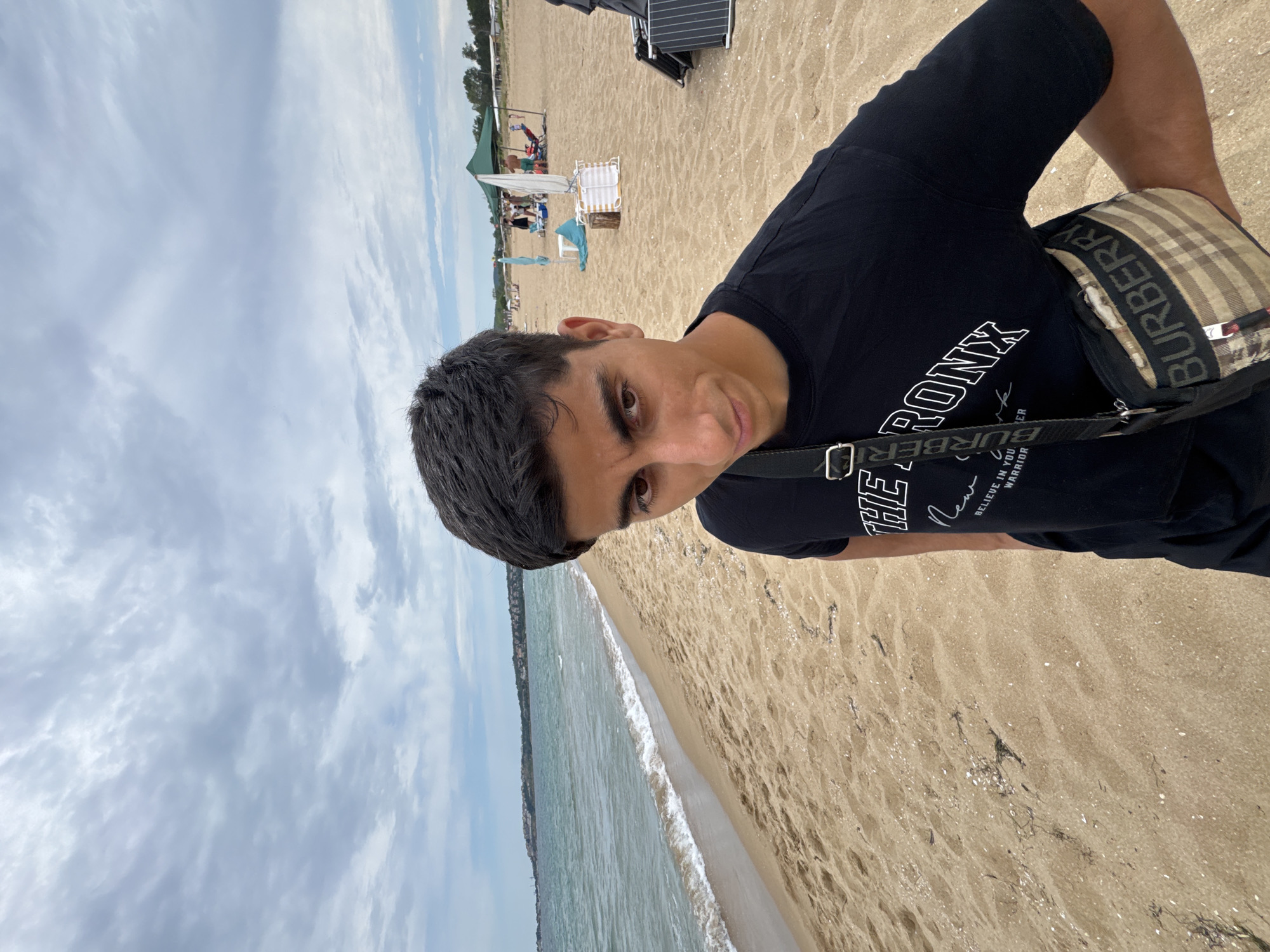 A young man, appearing to be in his late teens or early twenties, is captured in a selfie on a sandy beach. He has dark, short, styled hair and is looking directly at the camera with a neutral expression. He is wearing a black t-shirt with white lettering that reads "THE BRONX" and some smaller text below it, along with a crossbody bag featuring the Burberry check pattern and logo. The man is positioned on the left side of the frame.
The background reveals a beach scene characteristic of Sozopol, Bulgaria. The sandy shore stretches out, meeting a calm sea with gentle waves. The water is a greenish-blue color, and a distant coastline with buildings and greenery is visible on the horizon. The sky above is overcast with scattered clouds, suggesting a moderately bright day. Further down the beach, there are a few beach umbrellas, lounge chairs, and other beachgoers enjoying the day. The overall atmosphere is relaxed and typical of a summer day at a coastal resort.