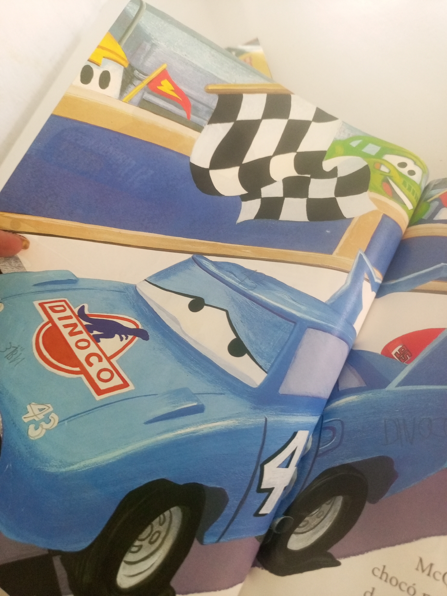 The image shows an open book with illustrations of cartoon cars. The left page features a blue race car with the Dinoco logo and the number "43" on its side. The right page shows another cartoon car, this one green, and a checkered flag. The book appears to be laid open on a surface. There are no people visible in the image, and the scene depicted in the illustrations is a racing environment. The background of the actual photograph is a plain, light-colored surface, possibly a table or a floor. The location in La Tagua, Venezuela is not discernible from the image itself.