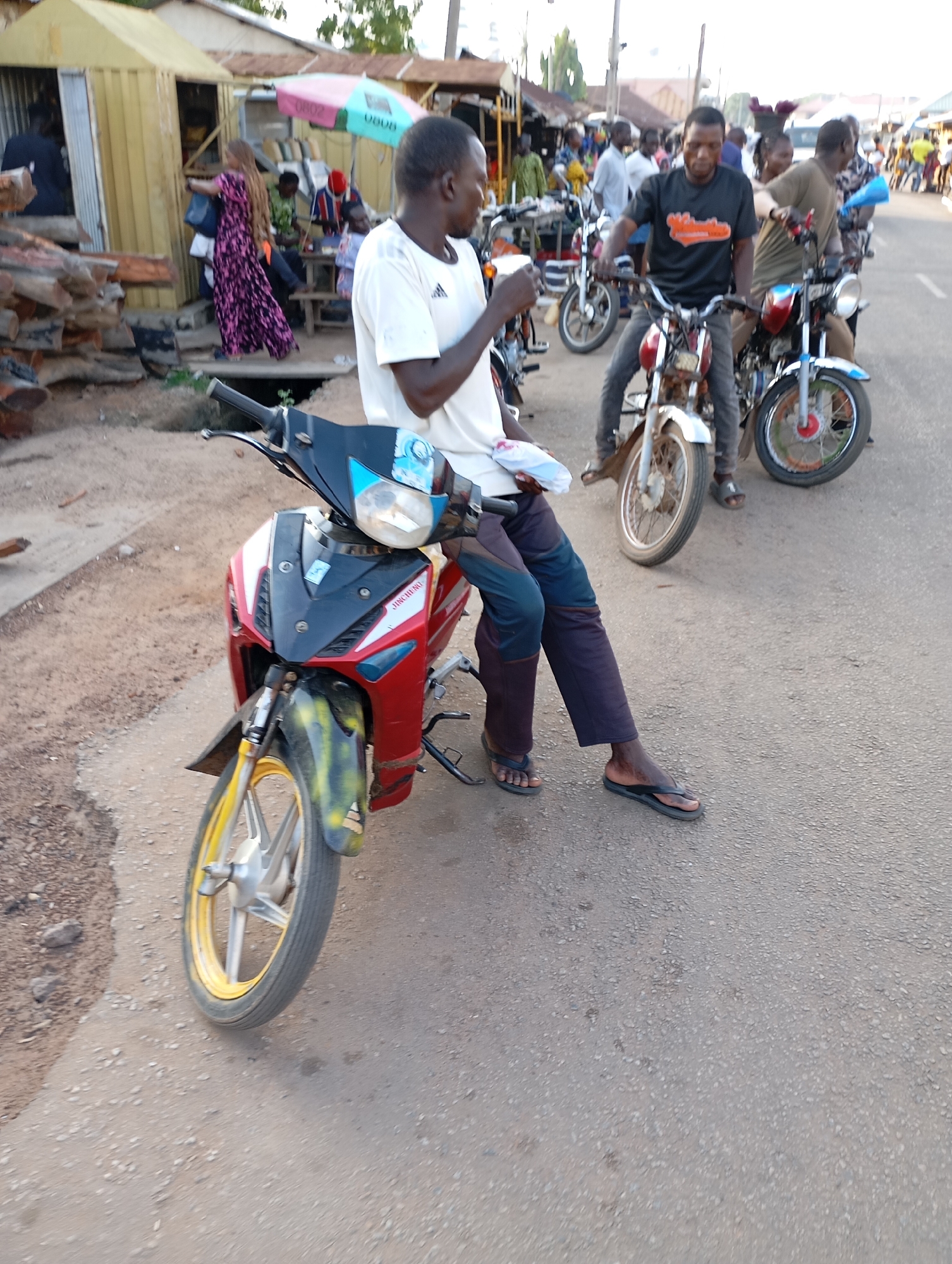 A medium shot shows a man sitting on a red and black motorcycle on a street in Shendam, Nigeria. The man is wearing a white t-shirt and dark pants. He is holding a white object in his right hand. The motorcycle is red with black accents. The front wheel of the motorcycle is yellow and black. In the background, there are other motorcycles and people. Some people are walking, and some are standing. There is a building on the left side of the image, and a street with a grey asphalt surface. The scene appears to be a market or a busy street.