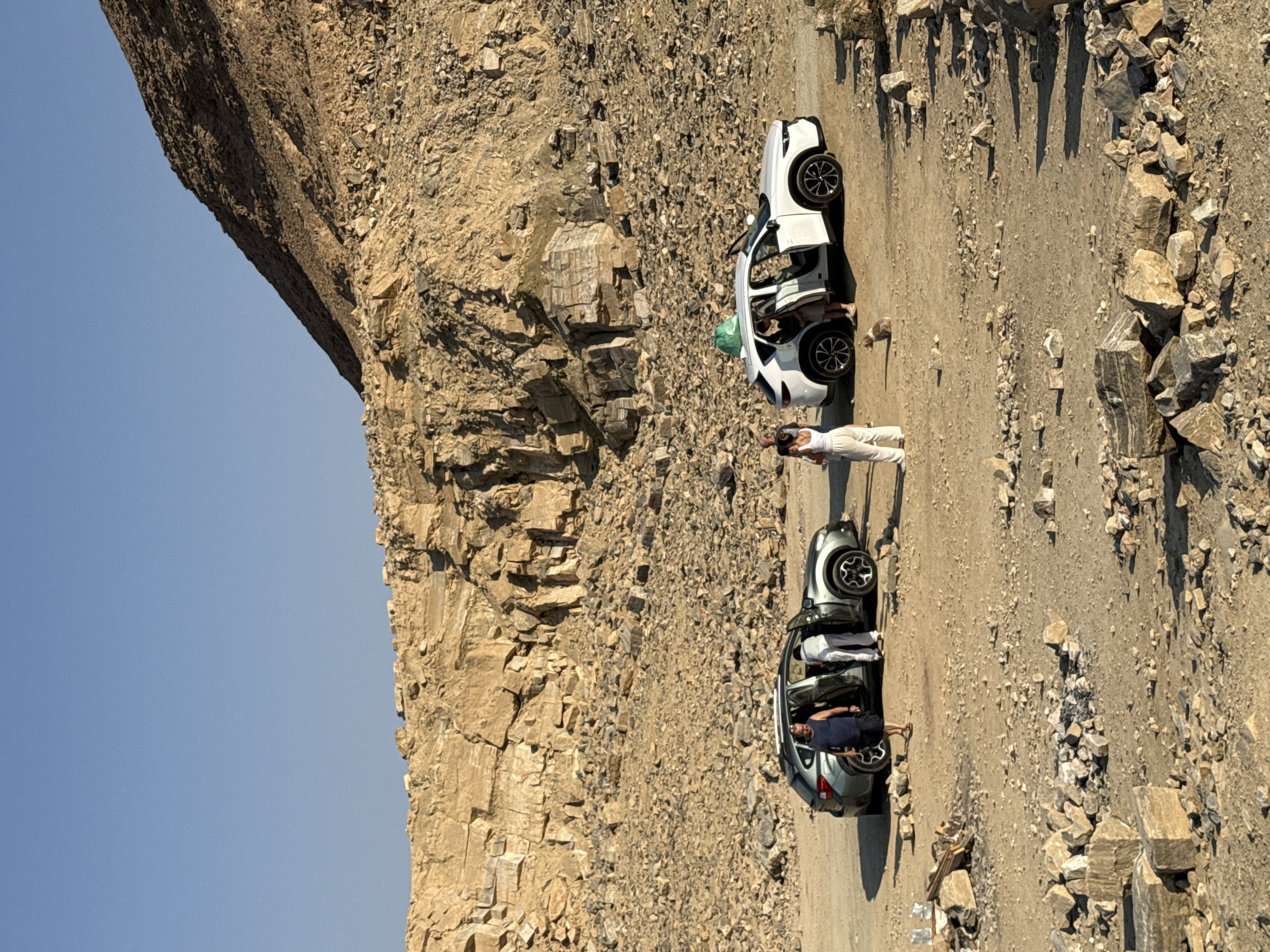 The image depicts a quarry in the Ramat Negev Regional Council, Israel, with two SUVs parked on a dirt road. The quarry has a large, rocky cliff face and is covered in loose stones and debris. A group of people are standing around the vehicles, some are getting in and out, and others are walking nearby. One SUV is a dark grey Subaru Outback, and the other is a white Mazda CX-5, which has a green bag on its roof. The sky is clear and blue, indicating a sunny day.