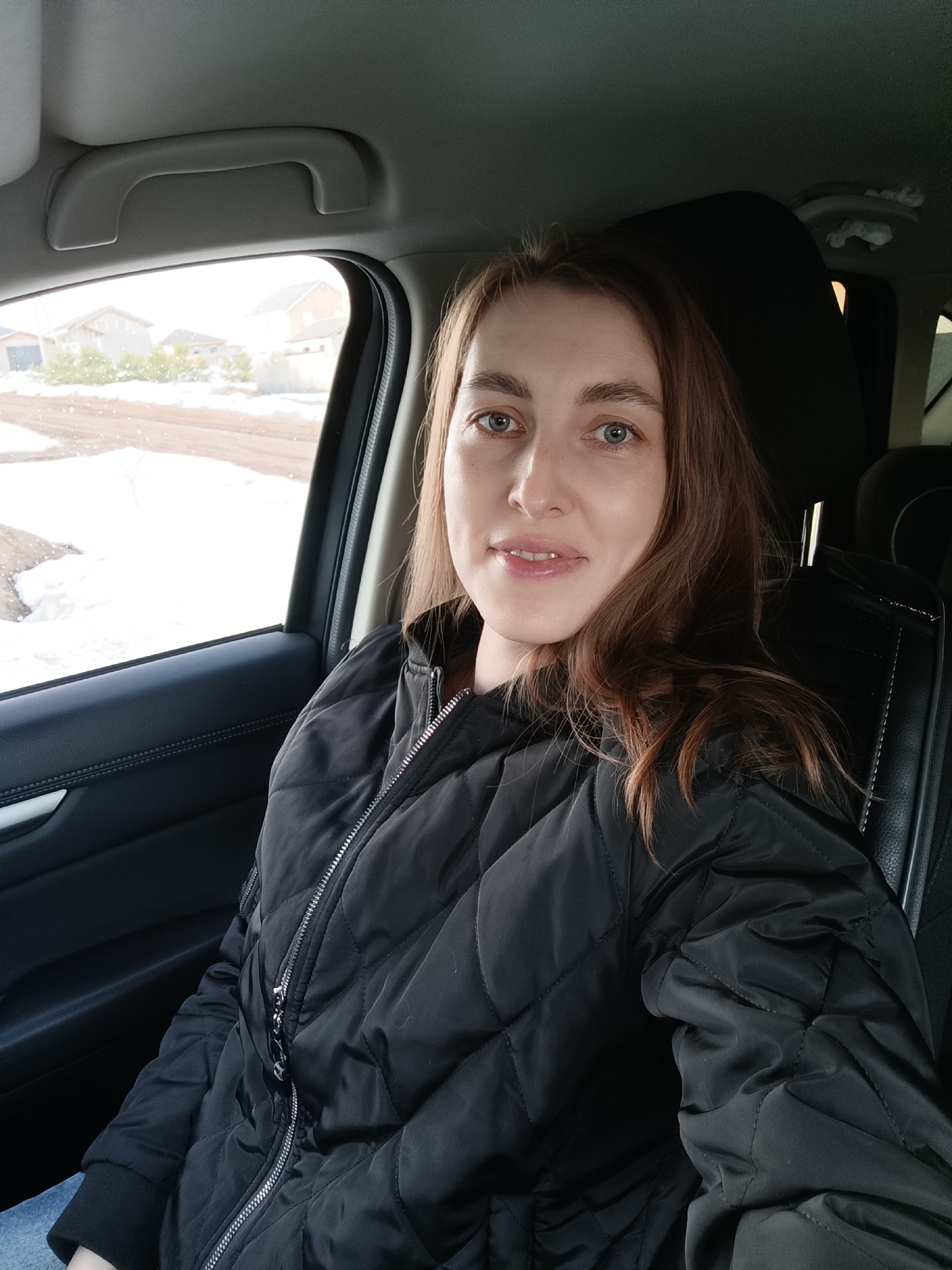 A woman is in the driver's seat of a car. She wears a black quilted jacket and blue jeans. Her hair is light brown and shoulder-length. Outside the car window, several houses and patches of snow are visible, suggesting the season is winter or early spring. The location is Chaikovsky, Russia.