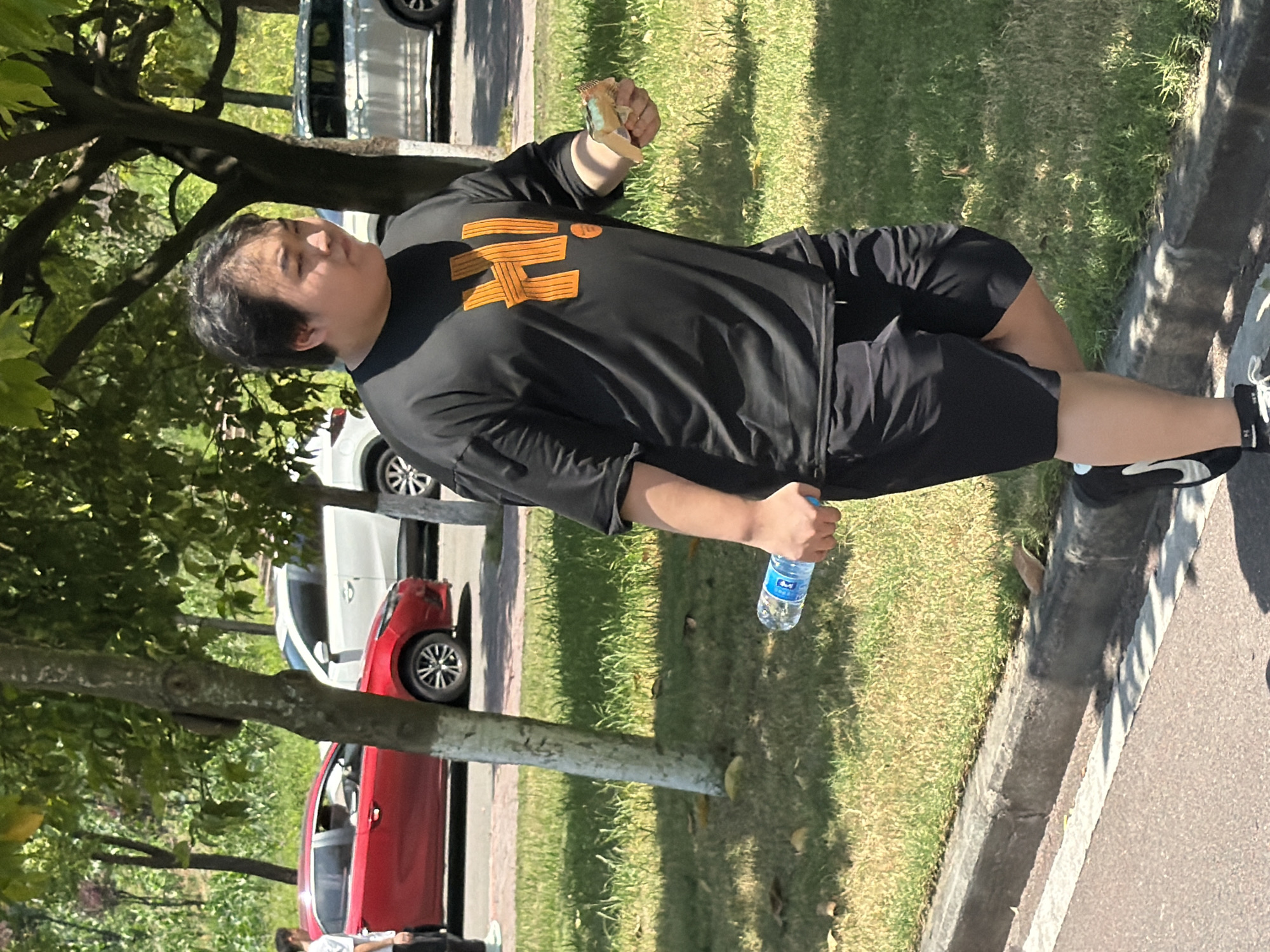 A person is walking on a paved path next to a grassy area with trees. The person is wearing a black t-shirt with an orange "Hi!" graphic and black shorts. They are holding a bottle of water in their left hand and a food item in their right hand. In the background, there are cars and trees. The scene appears to be outdoors in a park or urban area. The lighting suggests it is daytime.
