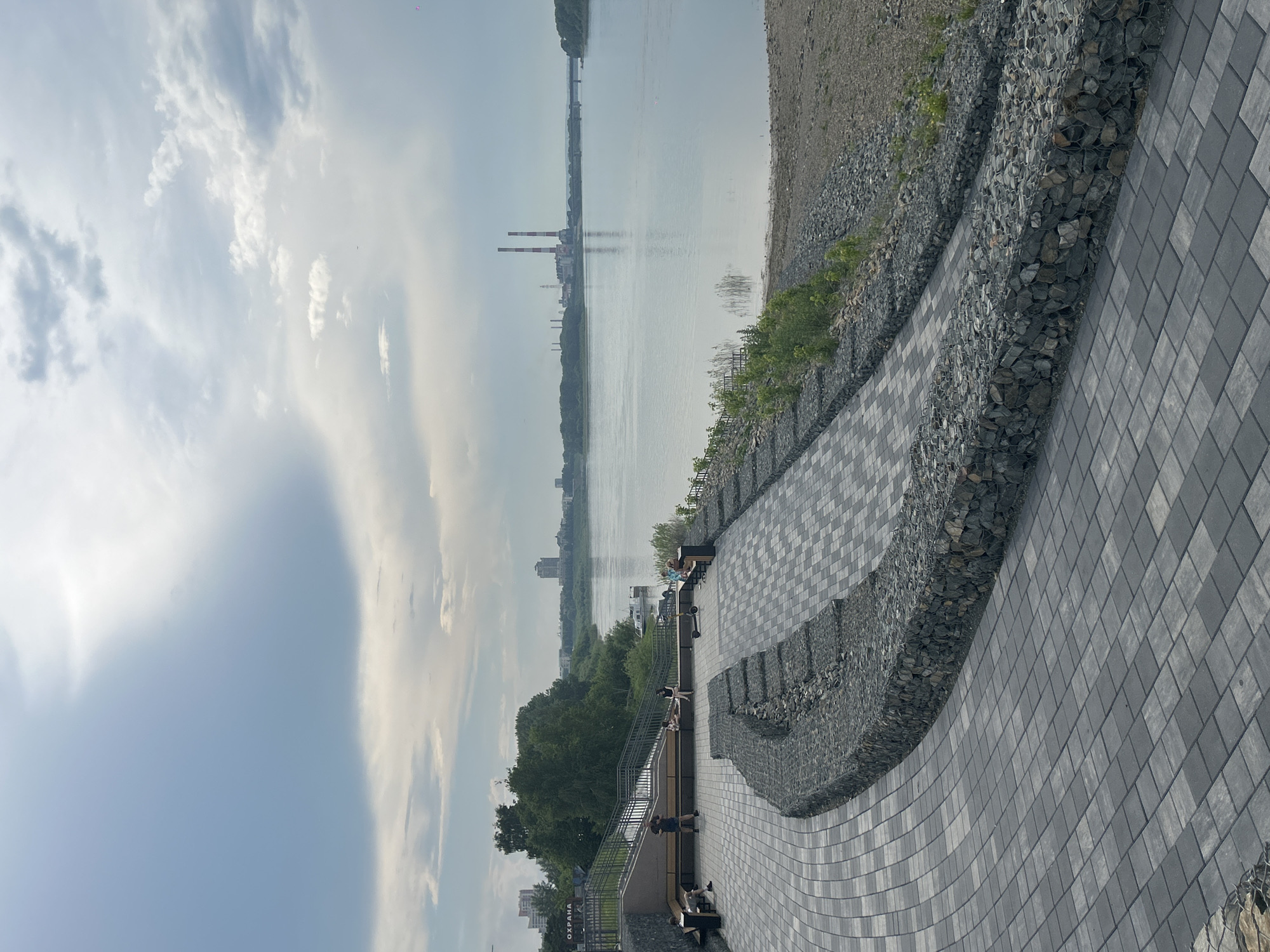 The image depicts a modern riverside promenade in Kemerovo, Russia, likely part of the Komsomolsky Park. The promenade features a winding paved walkway with a gabion wall on one side, filled with stones. On the other side of the walkway, a grassy area slopes down to a wide, calm river. In the background, across the river, are trees, buildings, and industrial structures, including prominent twin red and white smokestacks. Several people are visible on benches along the promenade, some sitting and others standing. One person is on an electric scooter. The sky is overcast with some patches of lighter clouds. The overall scene suggests a public recreational space by a river.