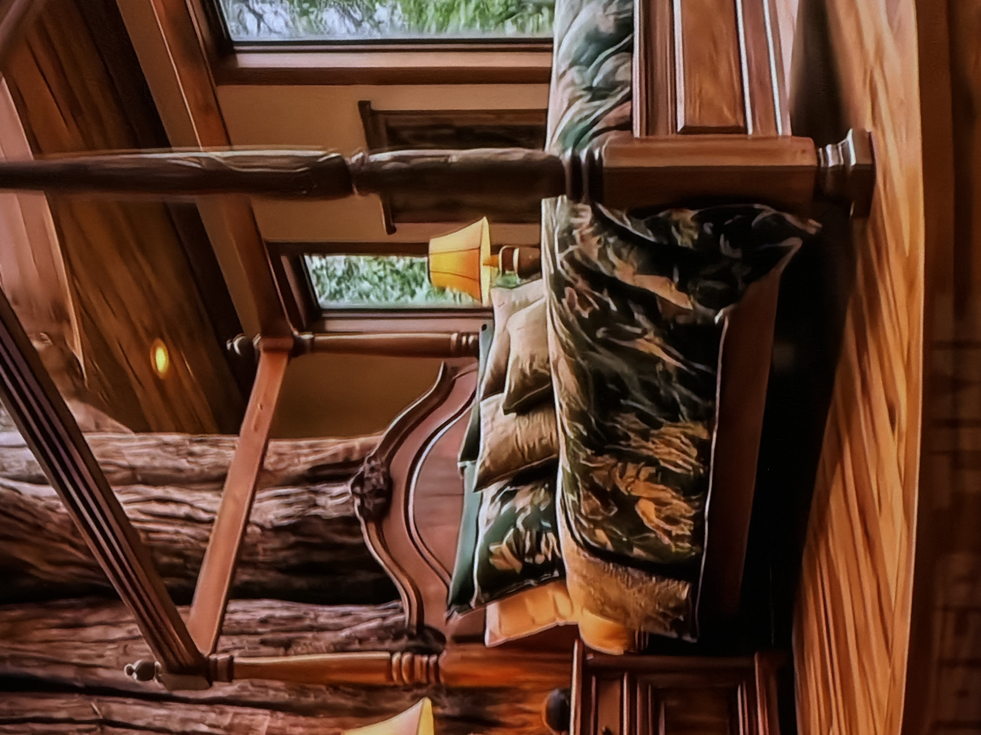 The image depicts a cozy, rustic bedroom, shown with a 90-degree clockwise rotation (described here as if upright). A prominent wooden four-poster bed dominates the scene, featuring large, dark wooden posts and a lighter wooden headboard with elegant carved details. The bed is generously appointed with numerous pillows and bedding in earthy tones, including shades of green, gold, and tan. Some of the fabrics display a camouflage-like or organic pattern.
Above the bed, windows or skylights reveal lush green foliage outside, suggesting a natural environment. A small lamp with a warm yellow lampshade is illuminated below the windows, adding to the room's inviting atmosphere. The interior architecture is characterized by substantial wooden elements, including rustic logs forming walls or structural supports, and polished wooden beams around the windows. The overall aesthetic suggests a lodge, cabin, or treehouse setting.
No people are visible, and there is no specific activity taking place beyond the presentation of the room. The time of day appears to be daytime, with natural light entering through the windows, complemented by the glow of the lamp. The scene evokes feelings of comfort, warmth, and tranquility. No visible text is present in the image.
