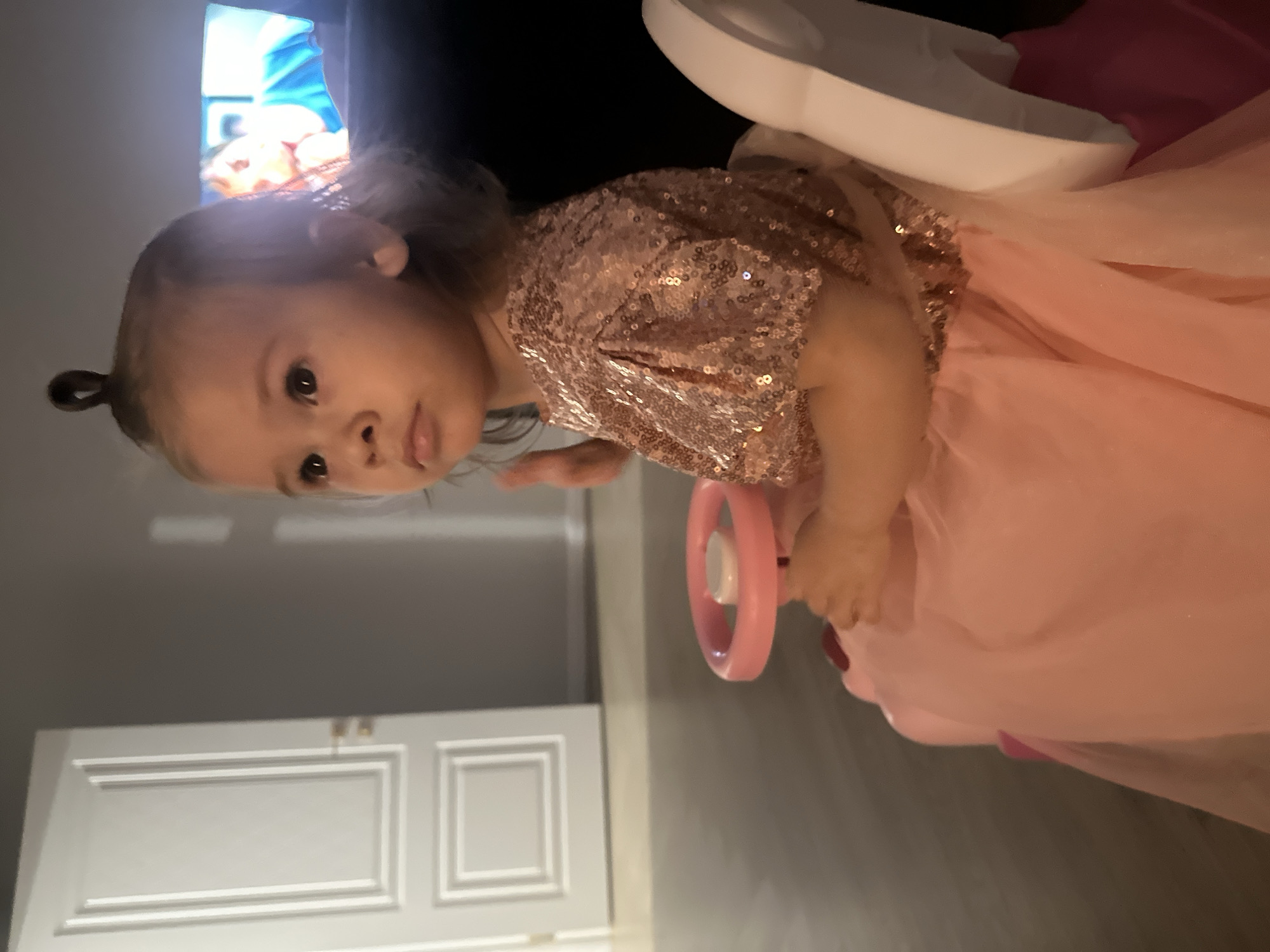 A young child with fair skin, styled hair, and wearing a shiny, sequined, light pink dress is sitting in a pink toy car indoors. The background shows a white door, a gray wall, and a glimpse of a television screen. The location in the city of Kharnavy, Russia is unknown.