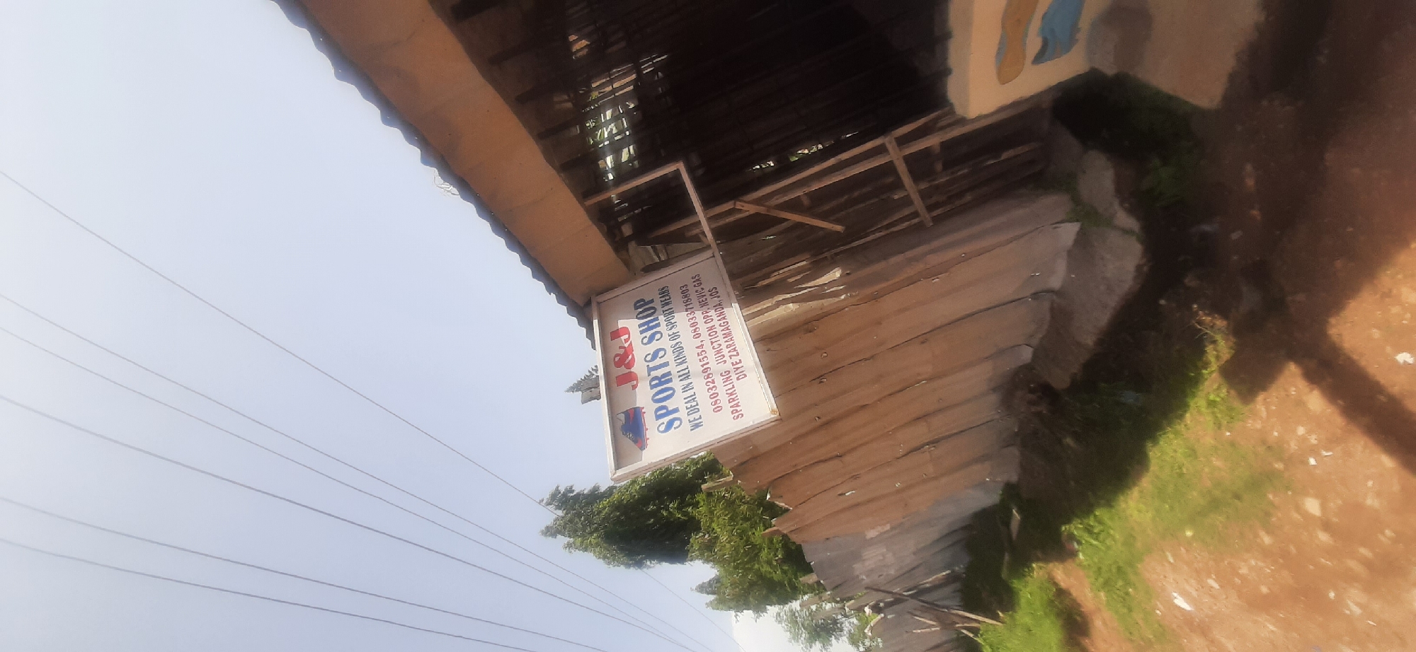 A sign for "J&J SPORTS SHOP" hangs on a wooden fence. The sign is rectangular with a white background and red and blue lettering. A graphic of a soccer cleat is visible on the upper left corner of the sign. Below the shop name, it states "WE DEAL IN ALL KINDS OF SPORT WEARS". Further down are phone numbers and an address: "08032891554, 08033718803", "SPARKLING JUNCTION OPP. NEVIS GAS", and "DIYE ZARAMAGANDA, JOS". The sign is tilted and partially obscured by the roofline of a building. The background shows a clear sky with visible power lines. The fence is made of corrugated metal sheets and is weathered. There is green grass and a dirt path in the foreground. The overall scene appears to be an outdoor commercial area in Jos, Nigeria.