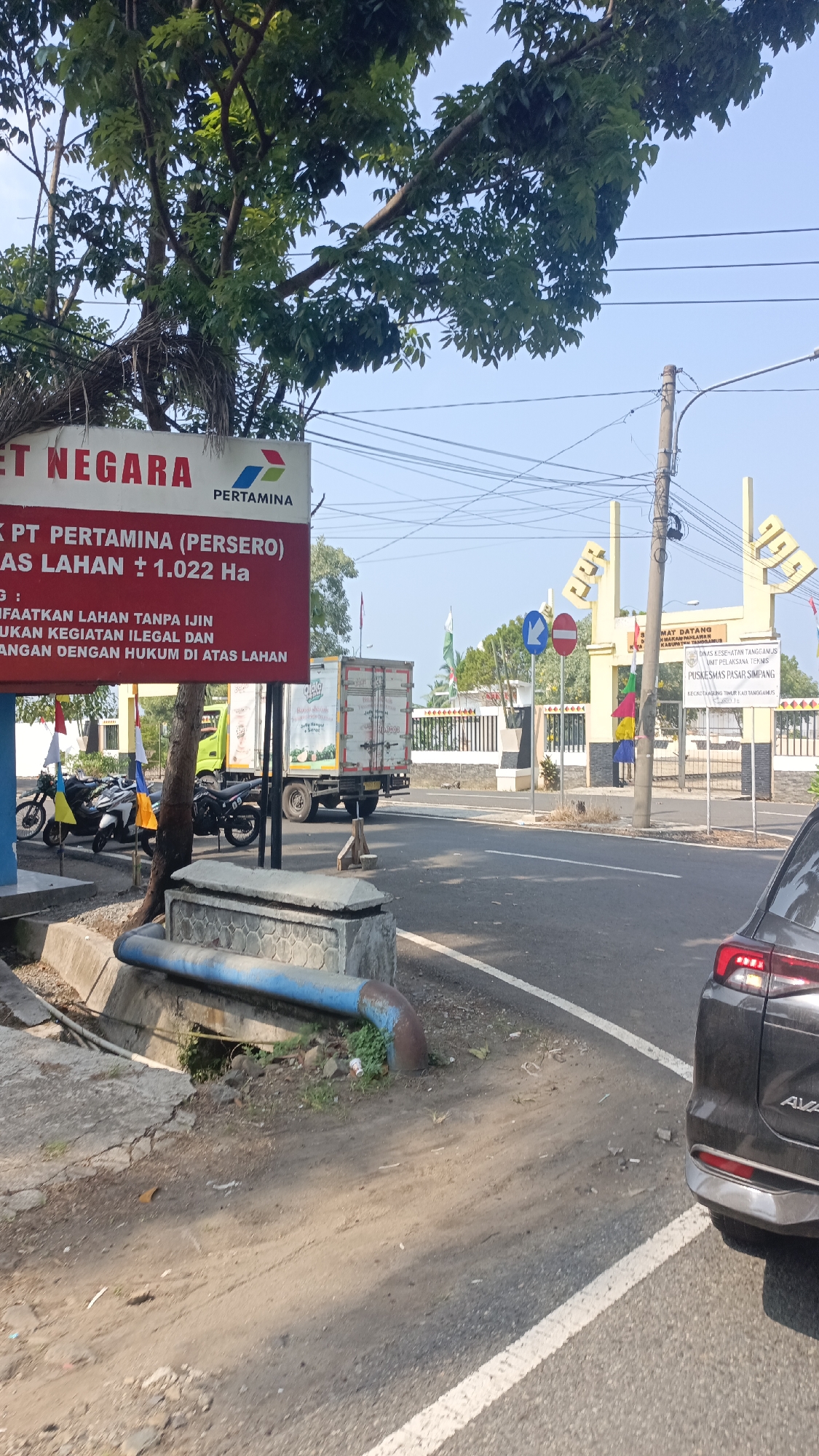 The image shows a street scene in Umbul Buah, Indonesia. A red sign with white and black lettering is prominent on the left side, displaying "ET NEGARA" and the "PERTAMINA" logo. Below this, text details PT PERTAMINA (PERSERO) land and regulations against illegal land use. In the background, a yellow gate with stylized text reading "CIGEBANG" stands, marking the entrance to a facility. Signs indicate "Dinas Kesehatan Tanggamus" and "Puskesmas Pasar Simpang." A truck is parked near the gate, and several motorcycles are lined up on the left. A dark-colored car is partially visible on the right. The environment appears to be a roadside with a clear blue sky overhead.