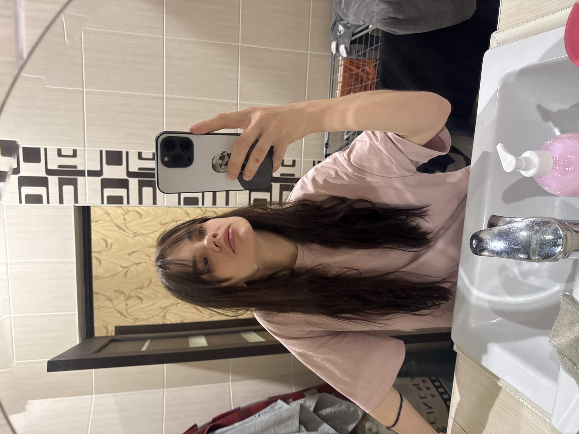 The image depicts a young woman taking a selfie in a bathroom. She is holding a smartphone with a light-colored case in her right hand, her reflection visible in the mirror. She has long, dark hair with bangs, and is wearing a light pink t-shirt.
The bathroom features a white sink with a chrome faucet and a pink soap dispenser. The walls are tiled, with a mix of light beige tiles and a patterned tile design behind the sink. A mirror is positioned above the sink, capturing the selfie. A glimpse of a laundry basket with various items is visible in the background.
While the image provides details of a domestic setting, no information within the photo suggests the location is Rostov-on-Don, Russia.