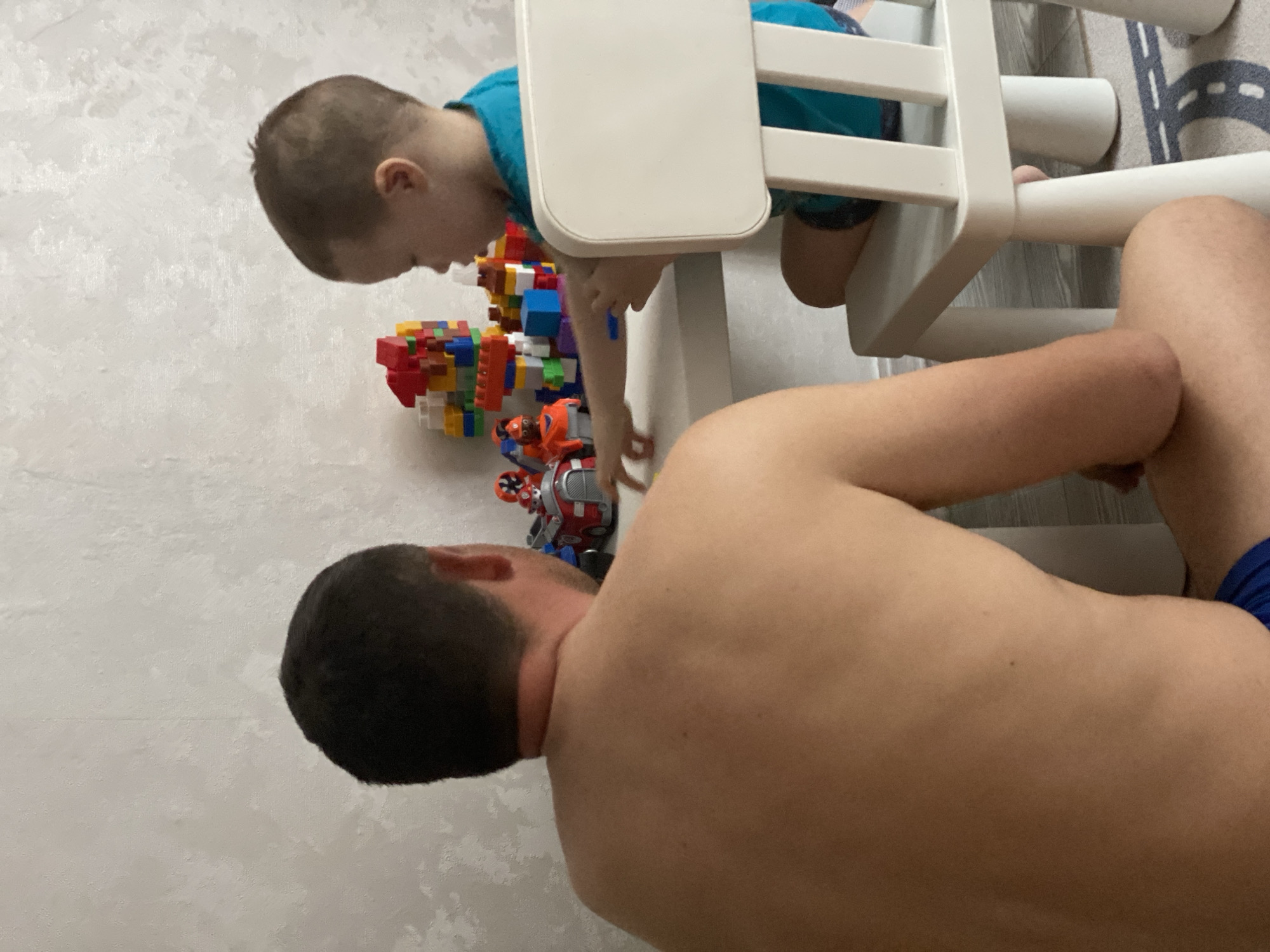A father and child are on the floor. The father is shirtless with dark hair. A child in a blue shirt is at a small white table, reaching for building blocks and toy figurines on the floor. They are in a room with a white wall, and a rug with grey, white, and black patterns. There is no indication that this photo was taken in Sochi, Russia.