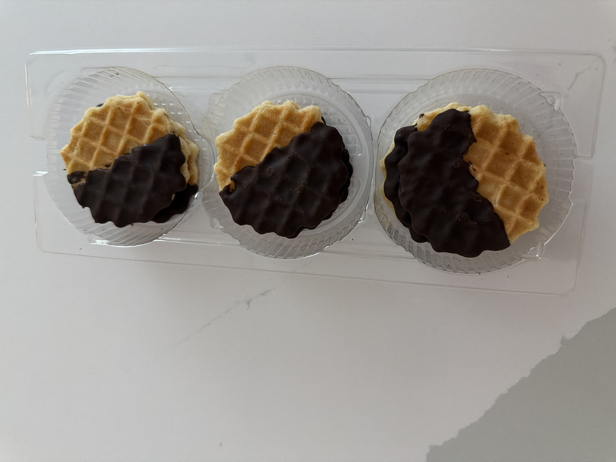 A top-down view shows three chocolate-dipped waffles in a clear plastic packaging. Each waffle is round with a diamond pattern on its surface and is partially covered in dark chocolate. The waffles are arranged vertically in a row within the plastic tray. The background is a plain, light-colored surface, likely a countertop. There are no people or animals present in the image. The scene appears to be indoors, possibly in a kitchen or retail setting. The lighting is soft and even, suggesting a well-lit room. There is a shadow cast on the left side of the image, obscuring a portion of the background. There is no discernible information in the image that would indicate a specific location in Chișinău, Moldova.