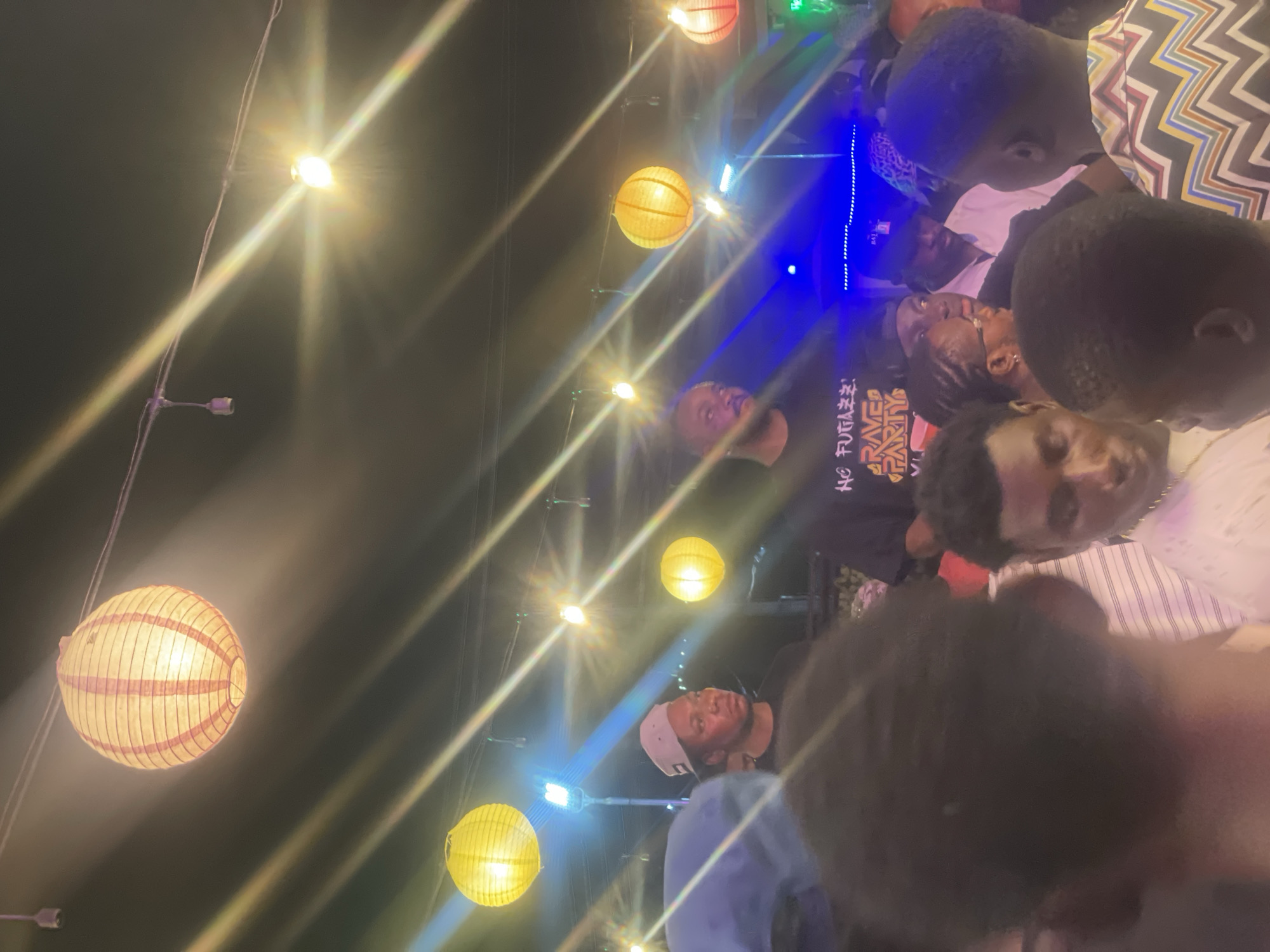 A nighttime scene in Lokoja, Nigeria depicts a gathering of people under illuminated decorative paper lanterns. The individuals are diverse in age and attire, some looking towards the camera while others are engaged in conversation. Overhead, string lights and colorful lanterns create a festive atmosphere. The location appears to be outdoors, likely a public space within the city.