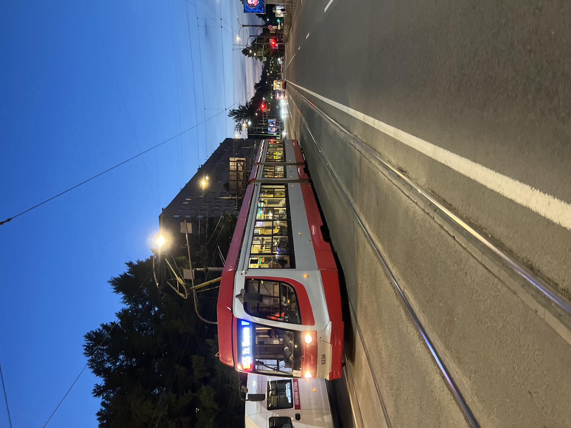 The image shows a red and white tram numbered 5236 moving along tram tracks on a city street at dusk. The tram is illuminated from within and has its headlights on. A white van is parked to the left of the tram. In the background, there are trees, buildings, streetlights, and traffic lights. The sky is a deep blue, indicating it is twilight. The tram is displaying "55 Ул. Воскова", suggesting it is route 55 going to Voskova Street. This location is consistent with Saint Petersburg, Russia. The scene is a street in a city with active public transportation and vehicular traffic.