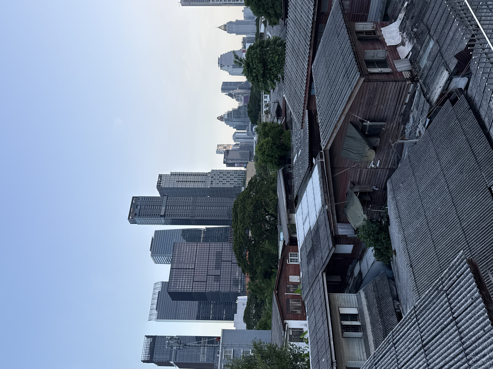 This is a high-angle, medium shot of a juxtaposition of old and new architecture in Bangkok, Thailand. In the foreground, a dense cluster of old, wooden homes with corrugated metal roofs, some showing signs of age and disrepair, are visible. Beyond these homes, a line of lush green trees separates them from a backdrop of modern, towering skyscrapers that dominate the city skyline. The sky is clear and pale blue, indicating it is likely early morning or late afternoon. There are no people or animals visible in the image. The overall impression is one of contrasting urban development within the city of Bangkok.
