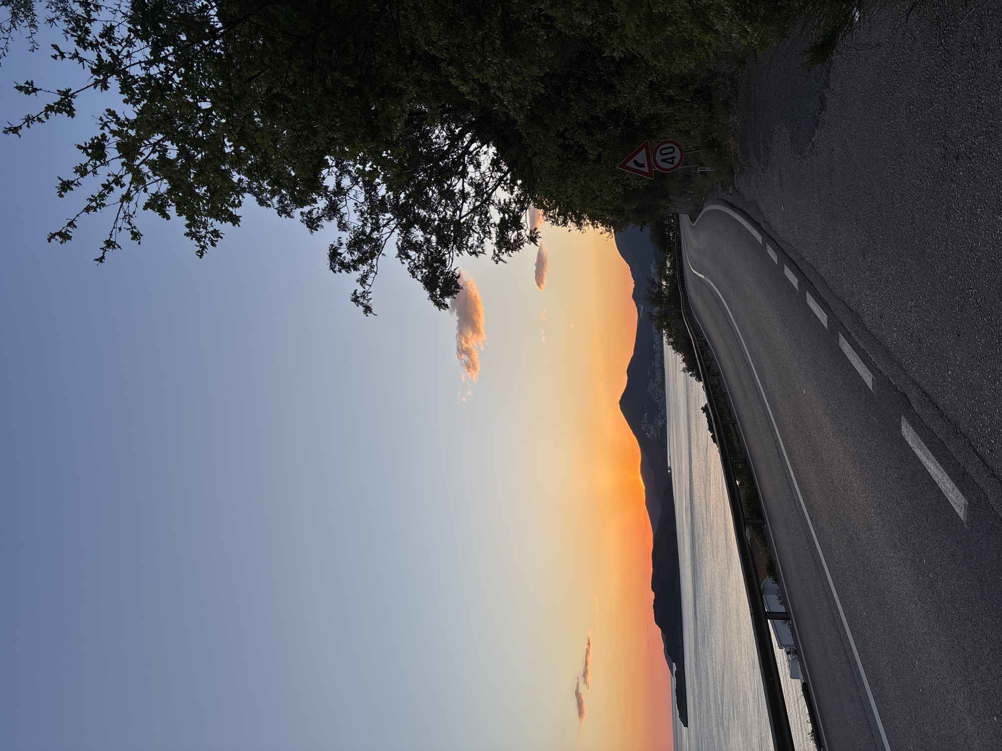 The image depicts a scenic view of a winding road along a body of water during either sunrise or sunset. The sky transitions from a soft blue at the top to vibrant orange and yellow hues near the horizon, indicative of the golden hour. Several small, wispy clouds are illuminated by the low sun.
In the foreground, a dark asphalt road with white dashed and solid lines curves to the right, following the contours of the land. A metal guardrail runs along the left edge of the road, separating it from the expanse of water, which reflects the warm sky colors.
Across the water, large mountains or hills are visible in silhouette, rising sharply against the brightly colored sky. Overhanging the scene from the upper left and top center is dense, dark green tree foliage, partially framing the sky and road.
Two European-style road signs are visible on the right side of the road, partially obscured by the foliage. One is a red triangular warning sign with an image of two consecutive curves, indicating a double bend ahead. Below or adjacent to it is a circular speed limit sign with a red border, displaying "40" in white on a black background, signifying a 40 km/h speed limit.
The scene conveys a sense of peacefulness and natural beauty, with no human activity or vehicles visible, focusing instead on the dramatic landscape and warm evening or morning light.