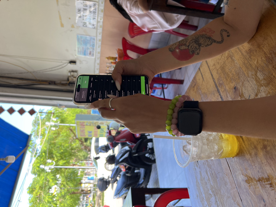 A person sits at a wooden table with a drink, looking at their phone. The phone displays a list of items. A smartwatch and colorful beaded bracelets adorn the person's wrist. A tattoo of a tiger sitting on a red ball is visible on their forearm. In the background, a street scene includes parked motorcycles and a business establishment with signs. Red plastic chairs surround the table.