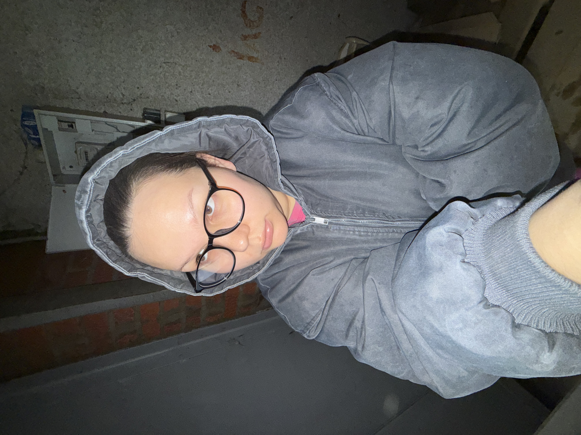 A medium close-up, portrait-style photograph shows a light-skinned person with dark hair wearing glasses, a pink shirt, and a gray hooded jacket with a zipper, apparently outdoors. The location shows possible brickwork, concrete surfaces, and what seems to be a white box of some sort. The location appears to be in the city of Chaikovsky, Russia. The individual looks toward the camera, and the image has a somewhat tilted orientation. The environment is dim and gives the impression of an urban setting.
