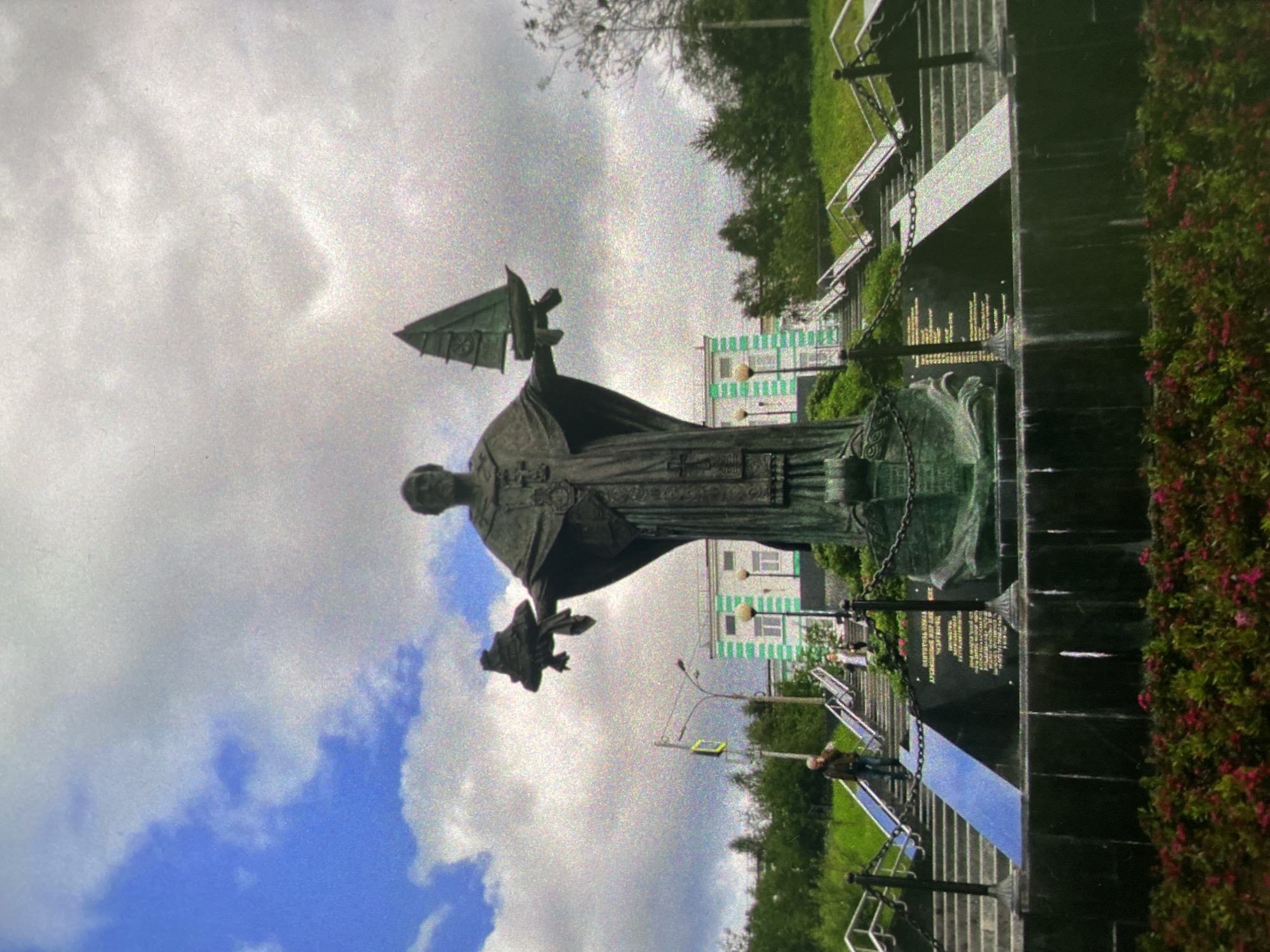 The image depicts a statue in Saint Petersburg, Russia. The statue is of a robed figure holding an object resembling a ship or flag. The statue sits atop a dark base, possibly granite, adorned with inscriptions. In the background, there are buildings with green and white facades, suggesting an urban environment. The sky is cloudy with patches of blue. A pedestrian is seen walking on a nearby pathway. There's greenery around the statue, including bushes with red flowers and grass. The setting appears to be a public space or park.