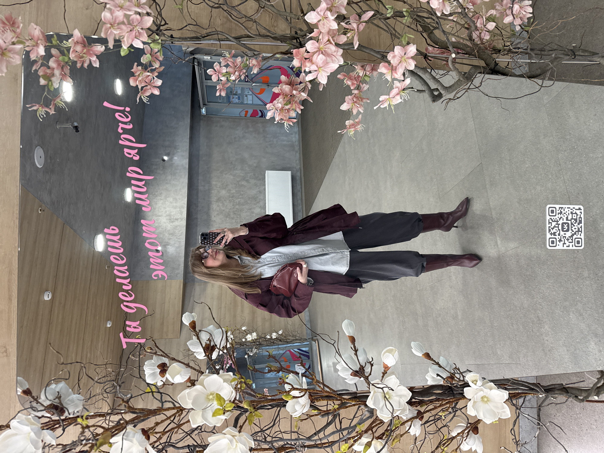 A woman is taking a selfie in a mirror decorated with floral arrangements. The mirror reflects an interior space with gray walls and floor. There are pink floral arrangements at the top of the mirror and white arrangements at the bottom. The woman is wearing a dark jacket, light shirt, and dark pants, and brown boots. She is holding a cell phone. The reflection also shows a QR code on the wall. In the upper background of the reflection, a doorway or window is visible. In the mirror, the text in pink is Russian, reading "Ты делаешь этот мир ярче!" which translates to "You make this world brighter!" A search reveals that the phrase and mirror decorations are located at the Izhevsk shopping center "Talismán" in Izhevsk, Russia.