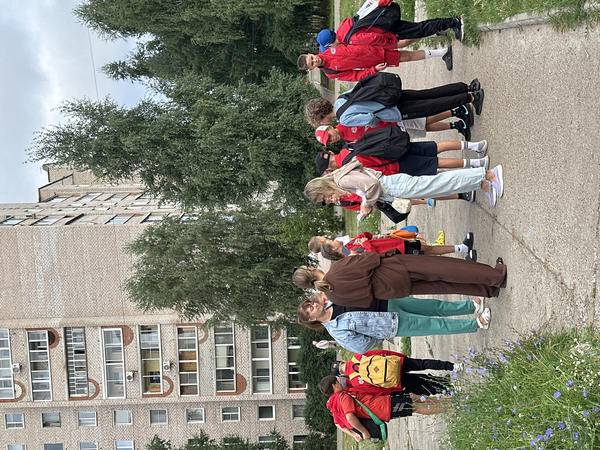 A group of children and adults, possibly a sports team, are gathered on a paved area in front of a multi-story brick apartment building. The children are mostly wearing red athletic clothing and backpacks. Two adult women are also present, one in a denim jacket and green pants, the other in a brown outfit. The environment appears to be outdoors, with trees and grass surrounding the paved area. A faint overcast sky is visible. The building itself has numerous windows and balconies, typical of Soviet-era apartment blocks. This scene is located in the city of Chaikovsky, Russia, an industrial city situated on the Kama River.